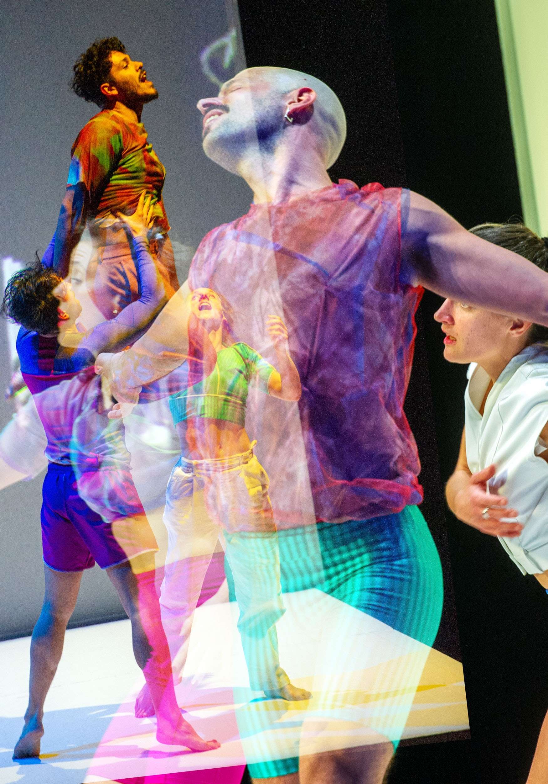L'image montre plusieurs danseuses et danseurs en pleine performance. On peut observer au moins quatre individus, bien que leurs images soient quelque peu superposées et diffuses, créant une impression de mouvement. La photo est très colorée avec des tons de rose, de bleu et de violet. 