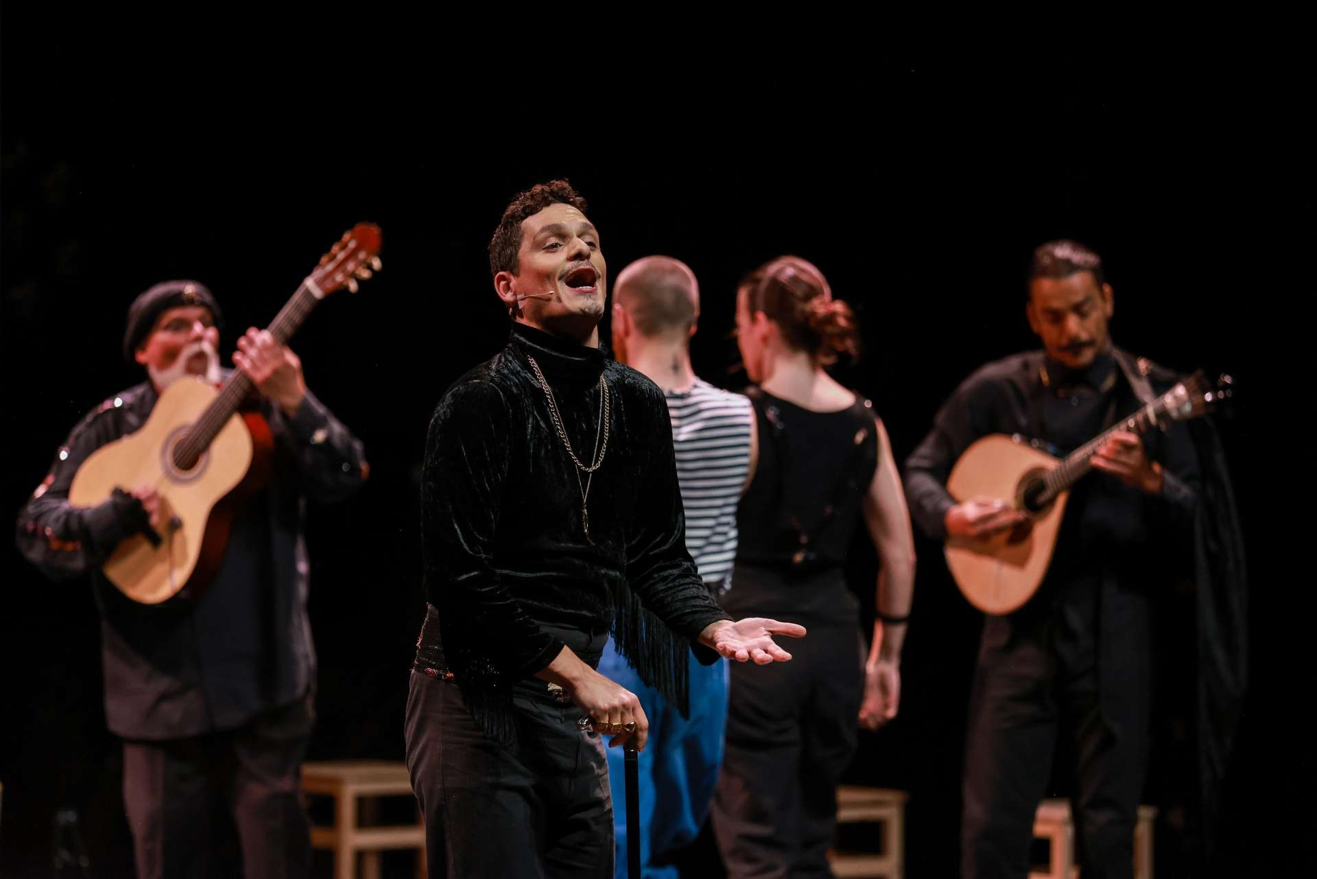 Chanteur de fado en premier plan, vêtu de noir, face au public. À l’arrière, musiciens, danseurs et danseuses également habillés de noir. En arrière plan un décor tout noir.