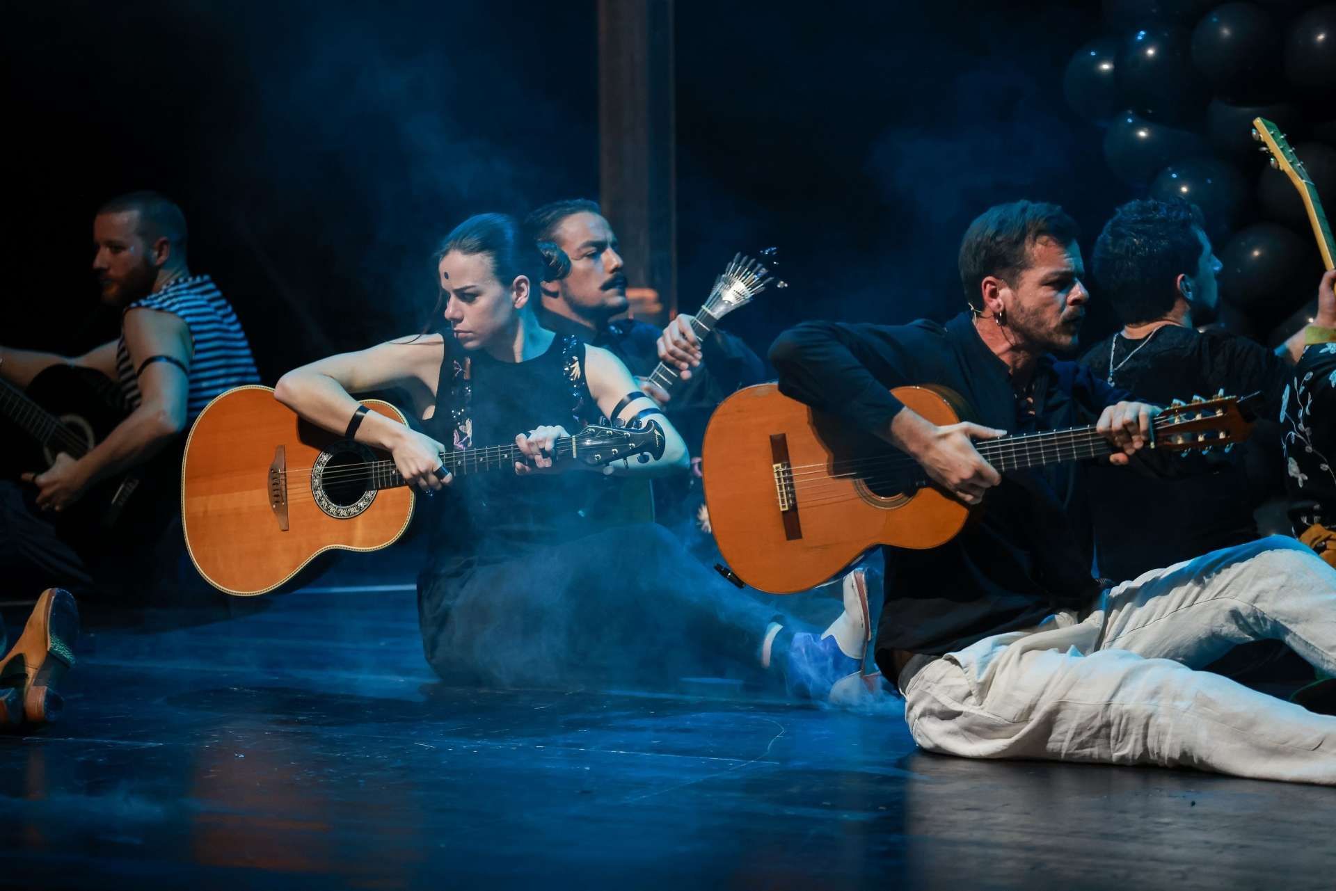 Danseuses et danseurs de fado, assis sur le sol, guitare à la main. En arrière plan, on aperçoit un structure de ballons noirs.