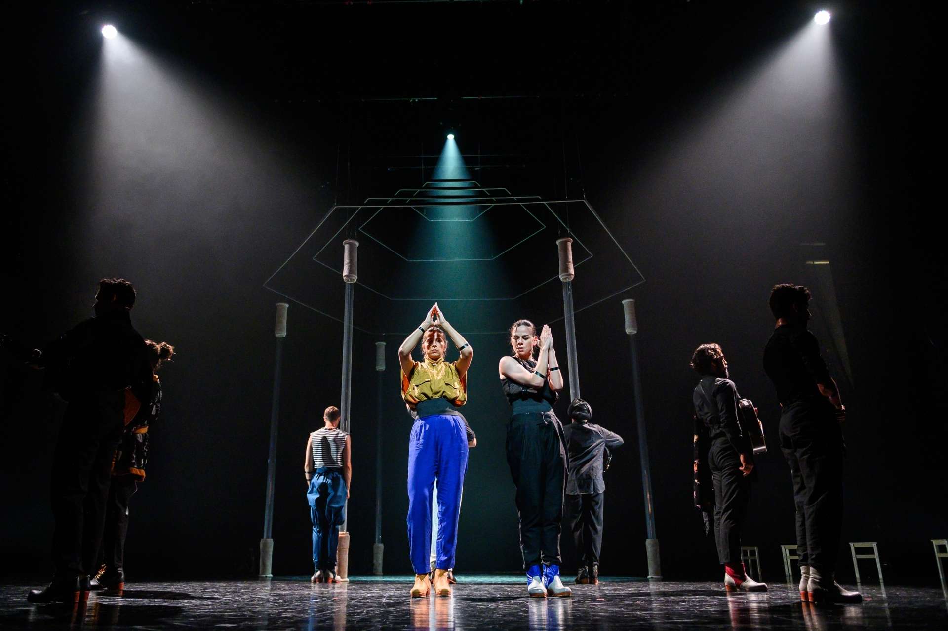 Deux danseuses sont positionnées au centre de la scène. Elles sont debout, bras levés et mains jointes devant elles dans un geste symétrique. Autour d’elles, six autres interprètes – musiciens et danseurs – sont répartis en arc de cercle. En arrière-plan, une structure suspendue composée de tubes métalliques forme une forme géométrique.