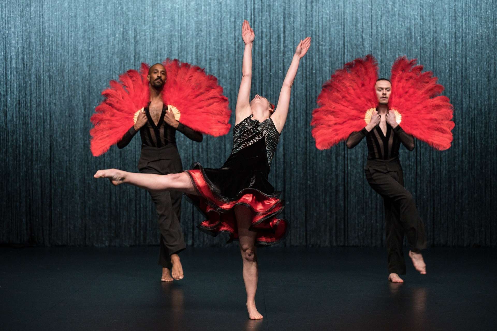 Trois danseurs sont le sujet principal de l'image. Ils sont vêtus de tenues noires. La danseuse du milieu porte une robe noir et rouge à volant en superposition, elle a les bras levés et une jambe perpendiculaire a son corps réalisant une pointe. Les deux danseurs derrière elle, portent de grands éventails en plumes rouges sur leurs épaules, dansent en synchronisation. 