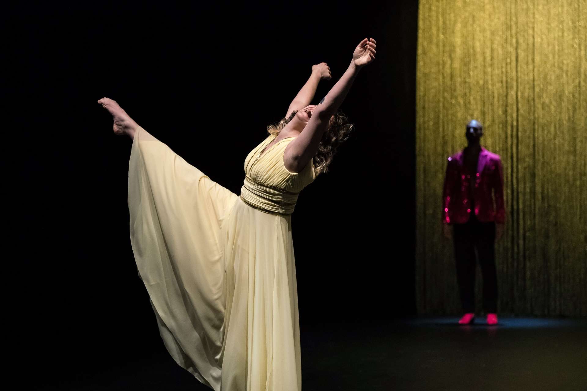 Photo d'une danseuse habillé d'une longue robe jaune pâle. Elle danse, les bras levés et fait un développé, sa jambe tendue et réalise une pointe. Derrière elle dans l'ombre on aperçoit un homme, surement un danseur, habillé de vêtement rouge à paillettes devant un rideau jaune. 