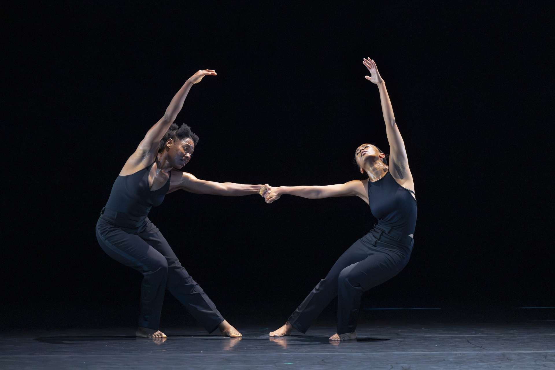 Photo de deux danseuses en pleine chorégraphie, elle se tiennent la bas pendant que leurs corps sont inclinés et tordus de manière harmonieuse, leurs bras gauches levés vers le ciel.