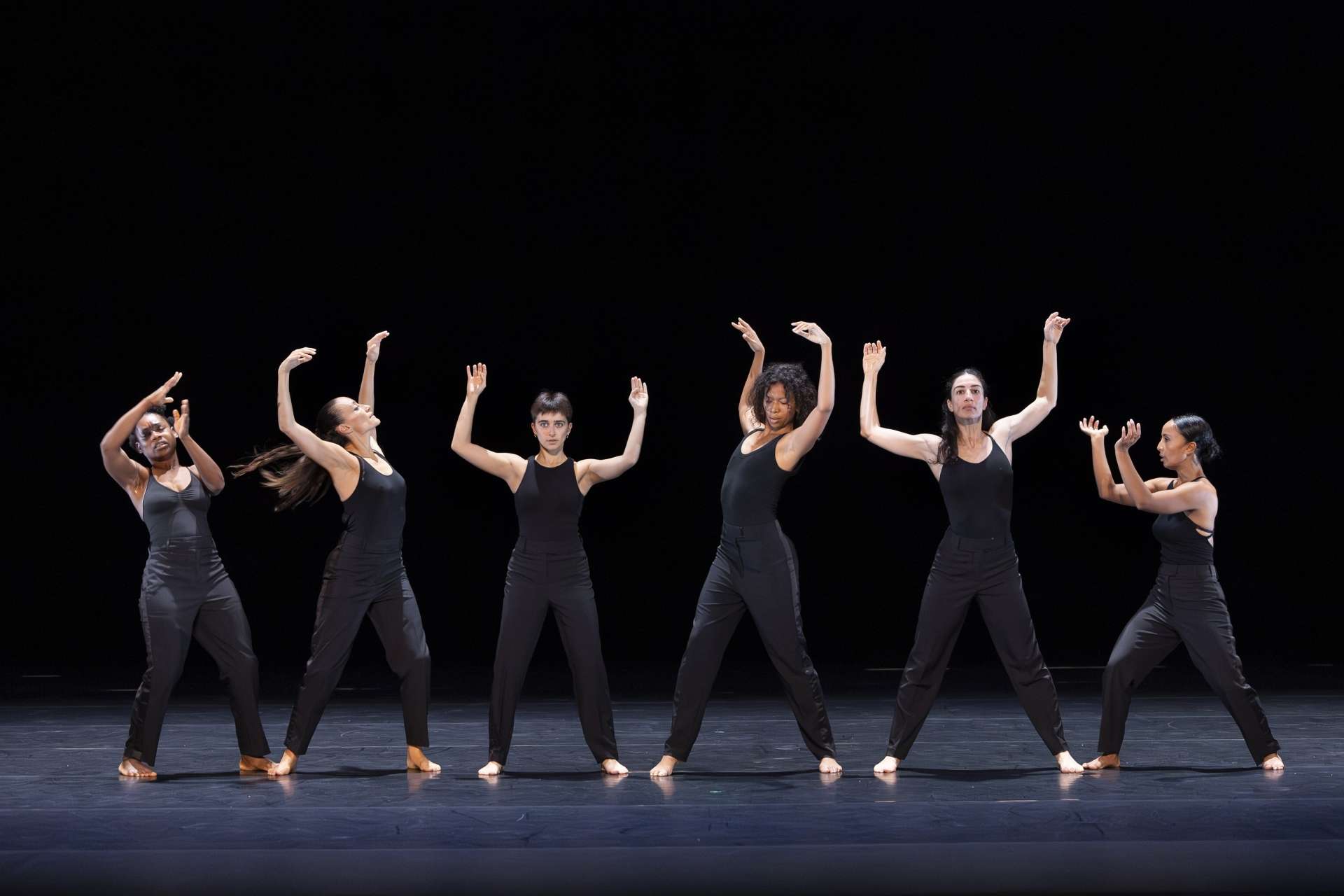 Photo d'un collectif de six danseuses, de face, vêtues de justaucorps noir qui exécute une chorégraphie de danse moderne jouant en synchronisation sur un fond noir. La chorégraphie met l'accent sur des mouvements de bras fluides et étendus vers le ciel. 