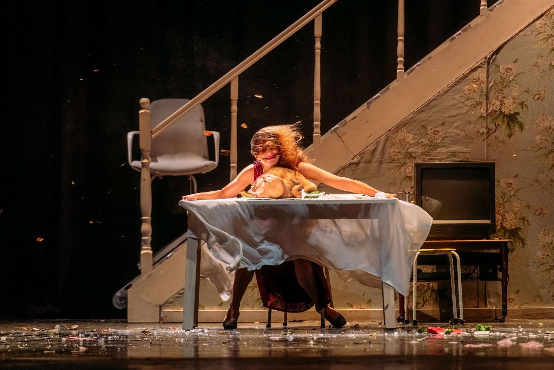 Photo du spectacle "Dimanche". Une femme assise sur une chaise est à table et se fatt emporter par le vent qui souffle dans la maison. Une dinde est dans un plat sur la table, et derrière elle on aperçoit des escaliers qui monte à l'étage. 