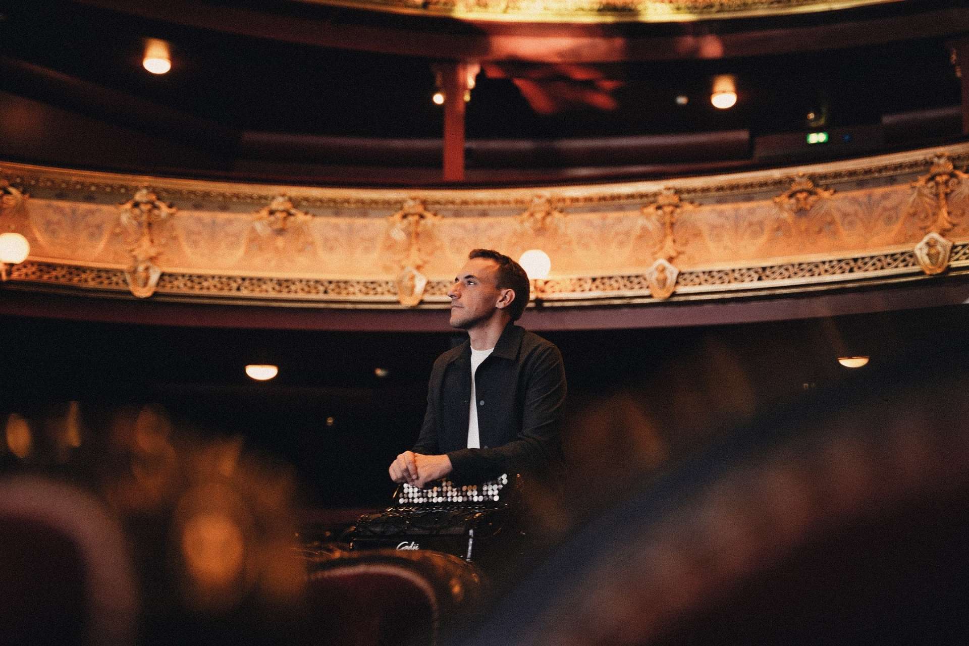 Photo de Félicien Brut, accordéon en main, au milieu d'un théâtre entres les sièges rouges. Premier plan et arrière plan flou qui laisse apercevoir l'architecture dorée de la salle. 