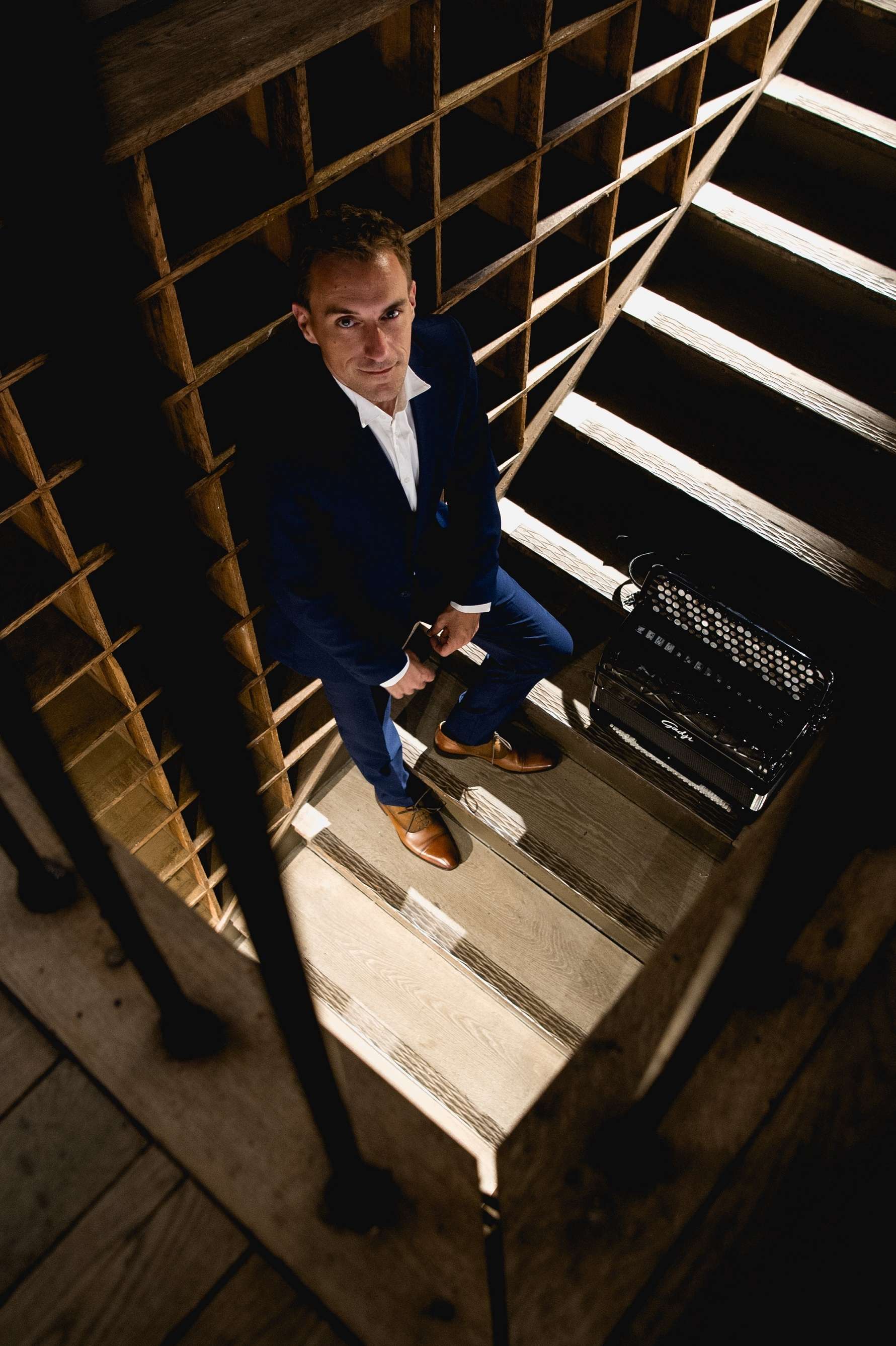 Photo de Félicien Brut dans les couloirs d'un monument. Il est débout et repose sur le mur à sa gauche, des escaliers montant à l'étage. Son accordéon posé sur le bas de ces escaliers. La structure est en bois.