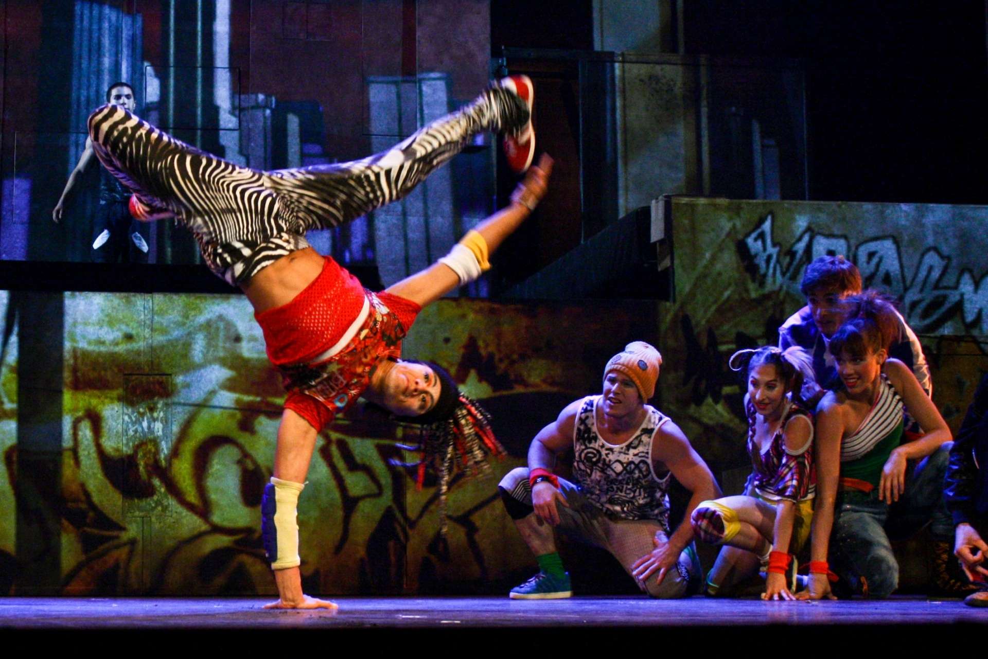 Placé au centre gauche de l’image, un danseur effectue une figure, (freeze ou handstand twist), typique du breakdance. À droite, un groupe, de danseuses et danseurs, est accroupi, observant ou réagissant à la scène.
