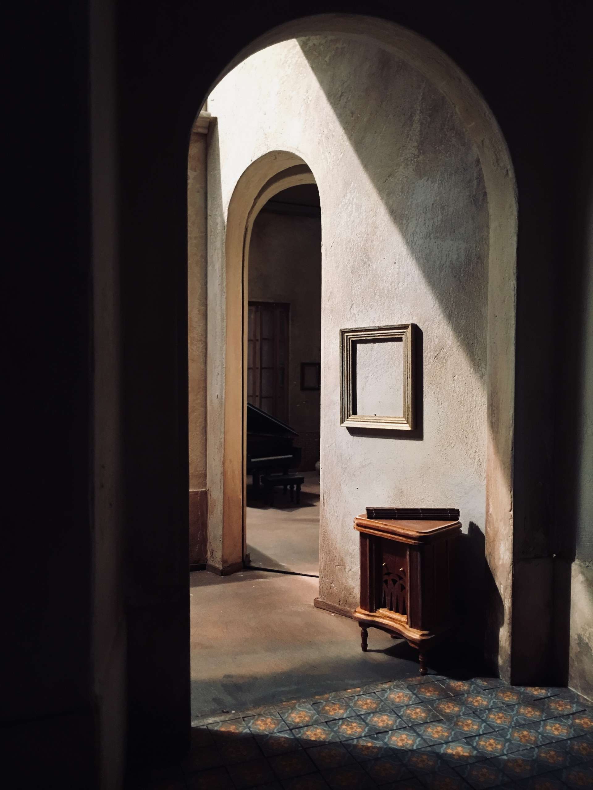 Image d'un couloir ou d'une pièce intérieure, avec des arches et un éclairage naturel contrasté. 3 sujets principaux dans l'image : un carrelage bleu avec des formes géométriques, un meuble triangulaire et un cadre vide fixé au mur. 