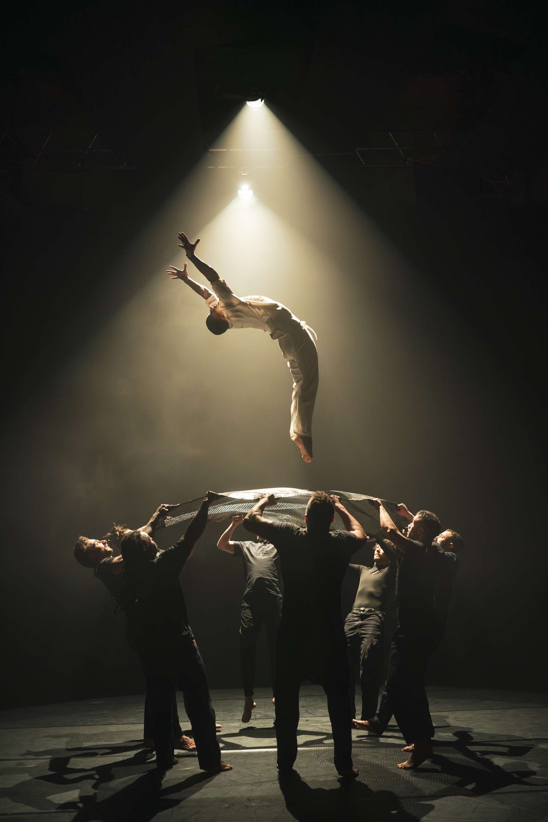 Un danseur, vêtu d'un costume blanc, est en plein saut dans les airs. En dessous, un groupe d'hommes, vêtus de tenues sombres, tendent un filet, leurs visages cachés par l'ombre. 