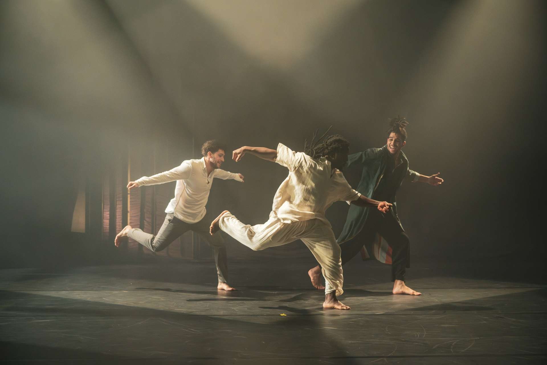 Trois danseurs pieds nus sont capturés en plein mouvement sur une scène sombre, éclairée par des projecteurs. 