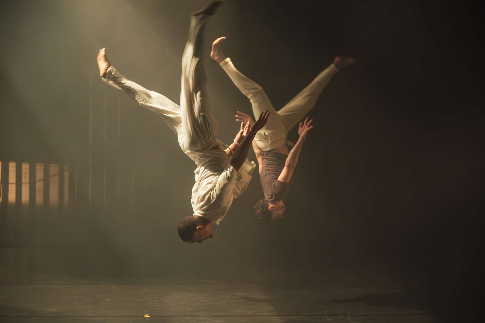 Deux danseurs habillé de vêtements amples et de couleurs neutres, font un salto synchronisé. En arrière une atmosphère remplie de fumée et un jeu de lumière.