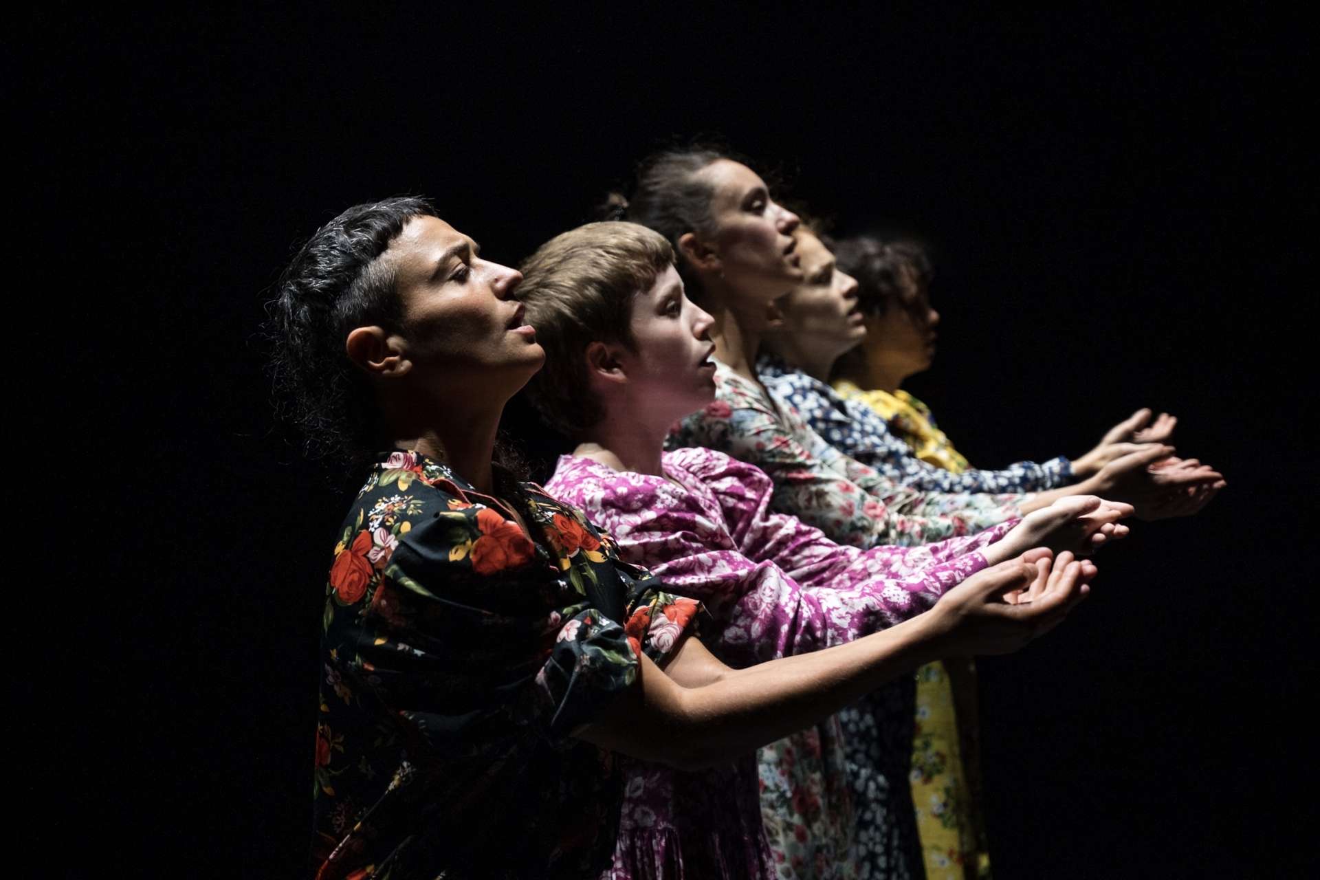 Cinque danseuses sont alignées et portent des robes à motifs différents. Elles sont de côtés, les bras tendus et les paumes des mains vers le ciel. En arrière plan, un fond tout noir.