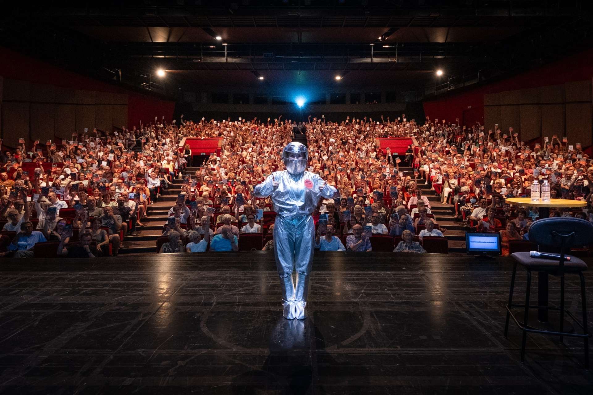 Sur scène, vue de la salle avec au premier plan Marie François (apprentie en communication) en tenue de cosmonaute et au second la salle comble avec les spectatrices et spectateurs qui lèvent leur brochure. 