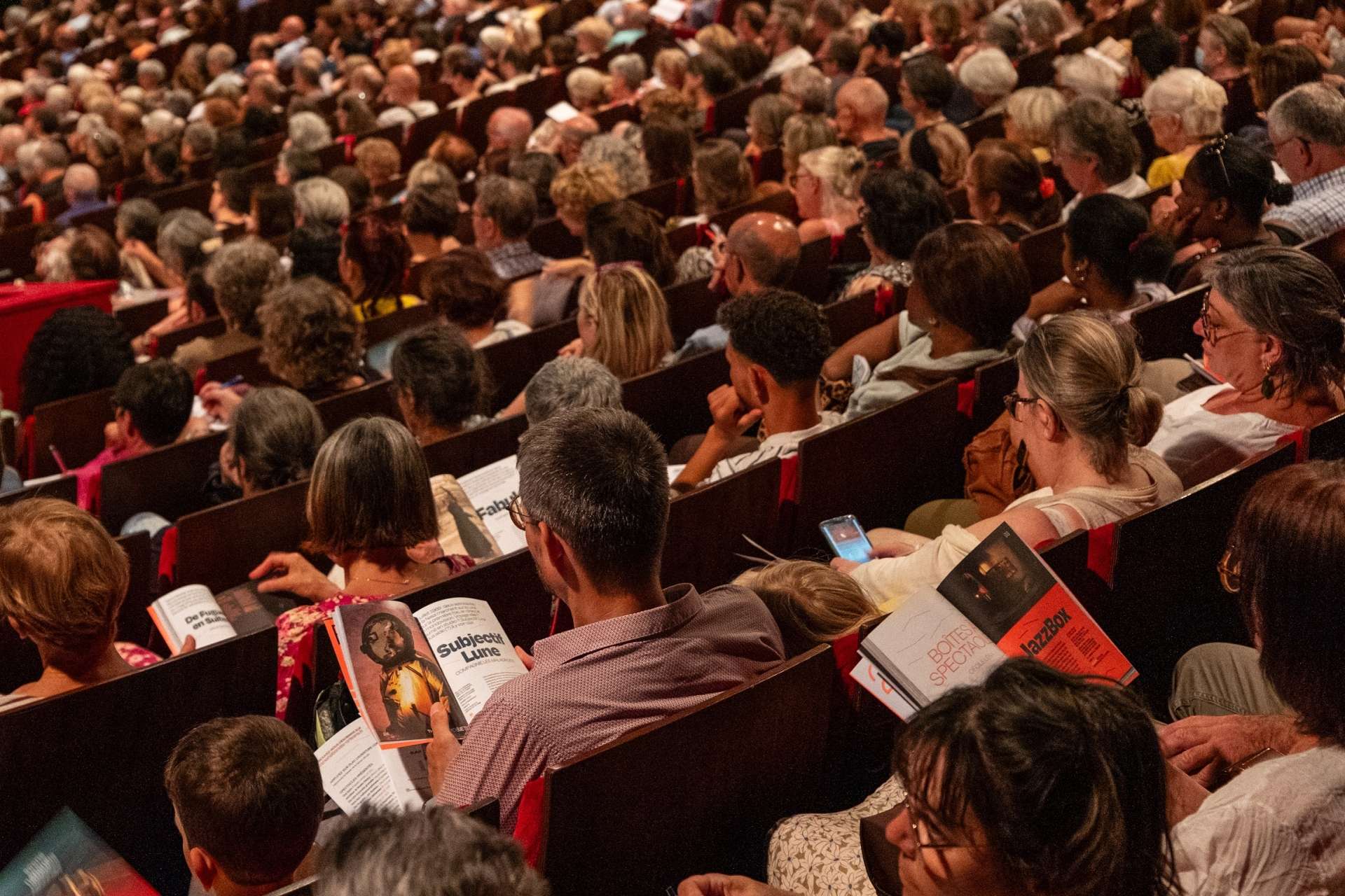 Photo du public de dos assis dans la salle Touchard, lors de la présentation de saison, feuilletant la brochure de saison 25/26 