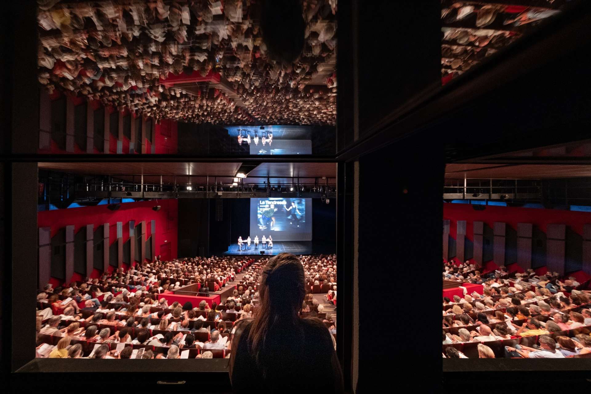 Photo de la salle Touchard comble prise de la régie. Une femme est installée au premier plan dans la pénombre et au-dessus d'elle, on peut observer le reflet de la salle. 