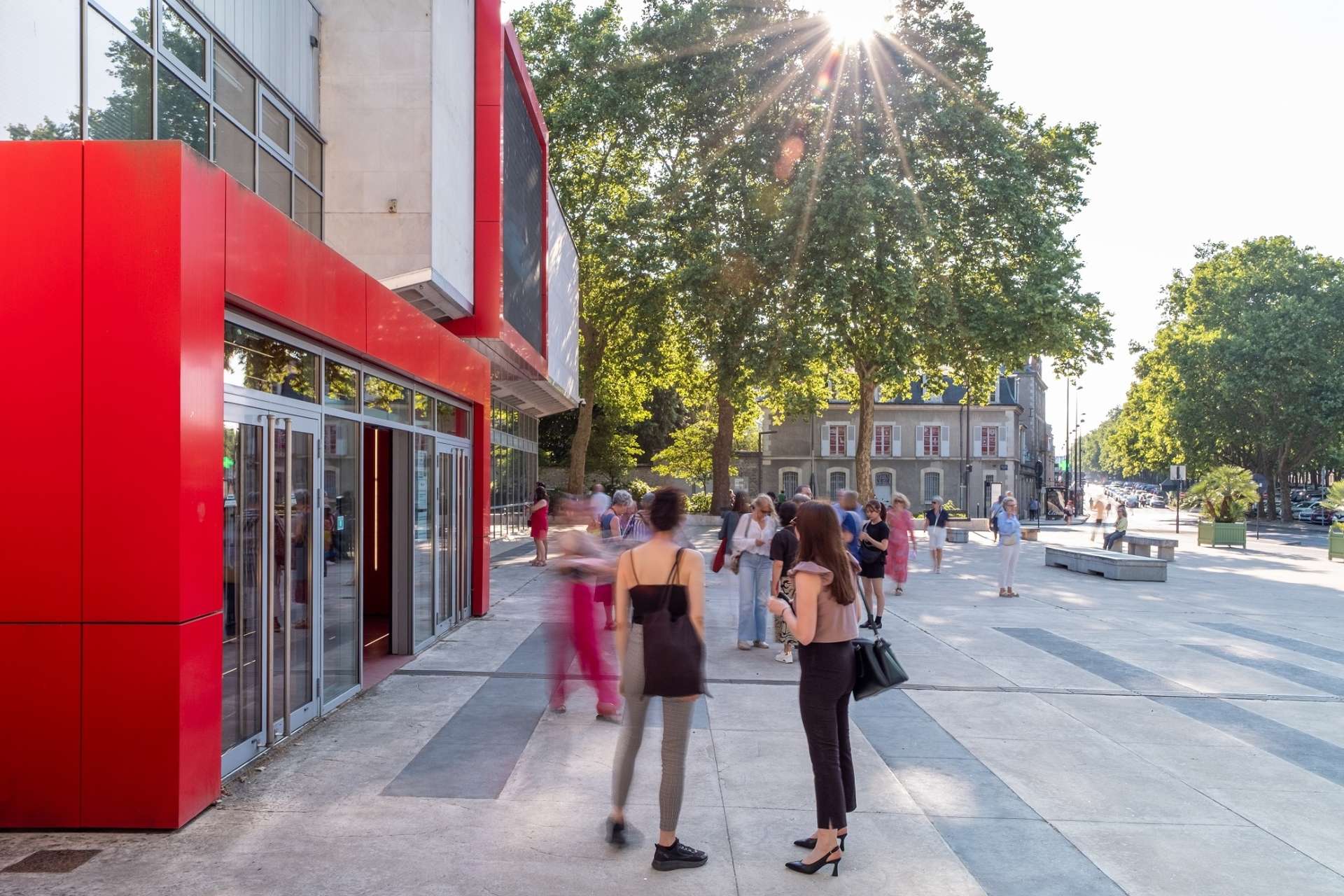 Vue de profil de l'entrée et du parvis du Théâtre d'Orléans. Du public est en train de rentrer par la grande porte entourée de rouge.