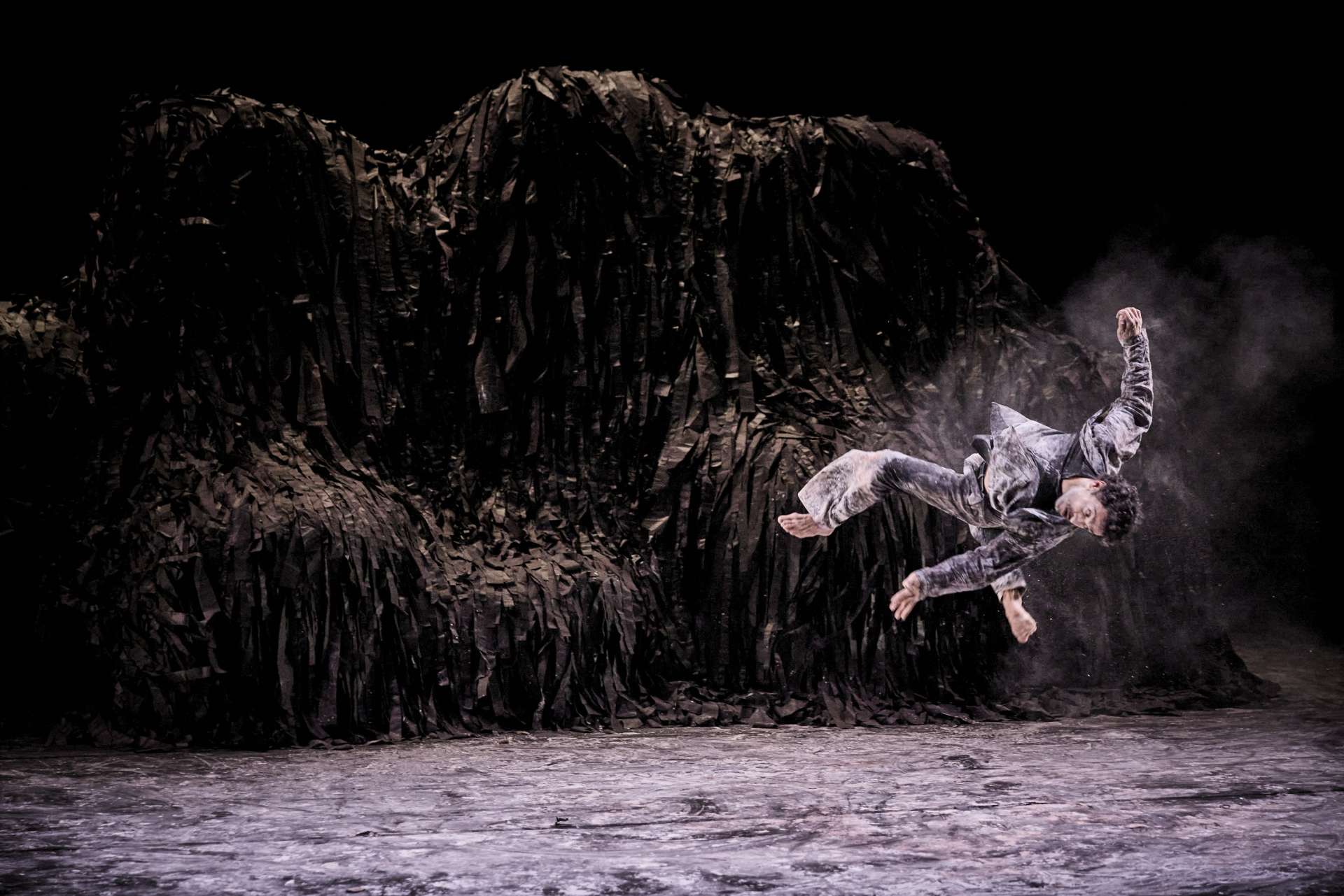 Photo d'un danseur en plein salto, projetant du sable de la scène autour de lui. Une structure ressemblant à une montagne, faite de morceaux de tissu noir est en arrière plan.