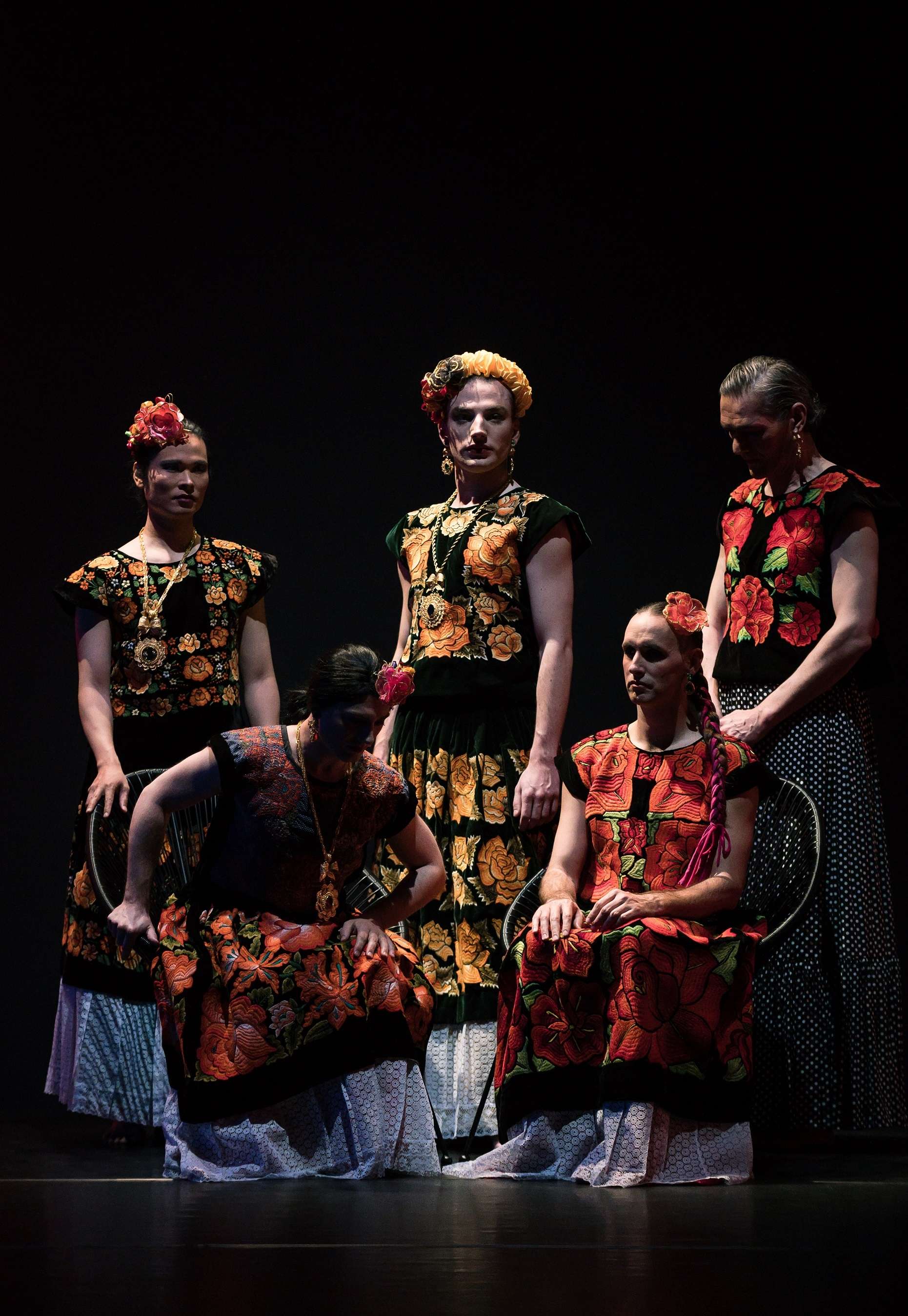 Cinq danseurs en robes fleuries et avec une couronnes de fleurs nous font face. Au premier plan, deux sont assis sur une chaise, tandis que trois autres se tiennent debout derrière eux.