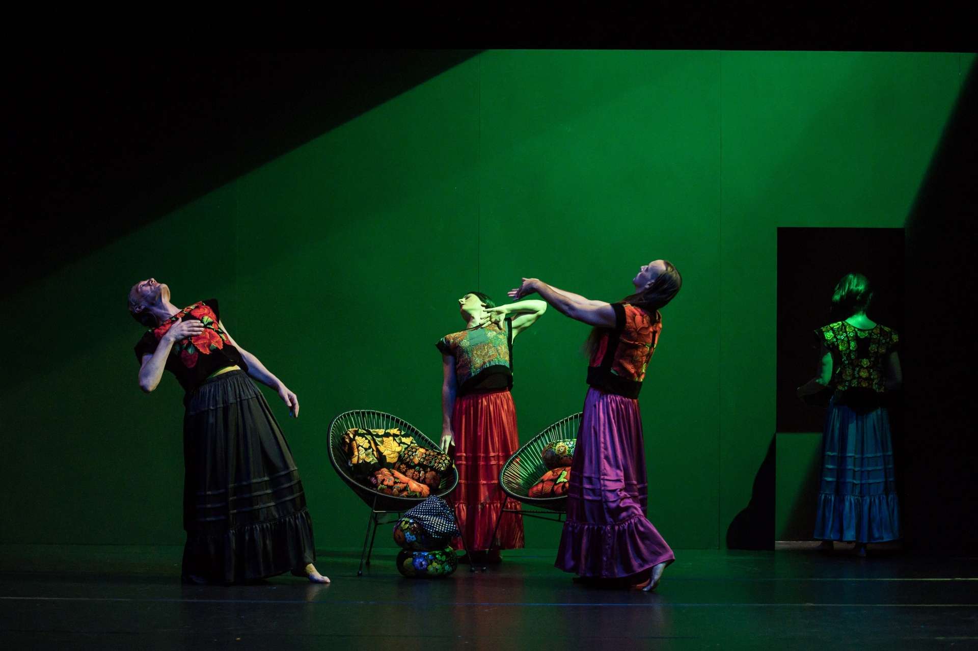 Trois danseurs portant des robes colorées (semblable à des vêtements traditionnels ou folkloriques) dansent autour de deux fauteuils, remplis de tissus ou de coussins à motifs colorés. Plus à droite et légèrement en retrait dans une zone plus sombre, un quatrième danseur, habillé lui aussi en robe, est en grande partie cachée mais visible de dos.