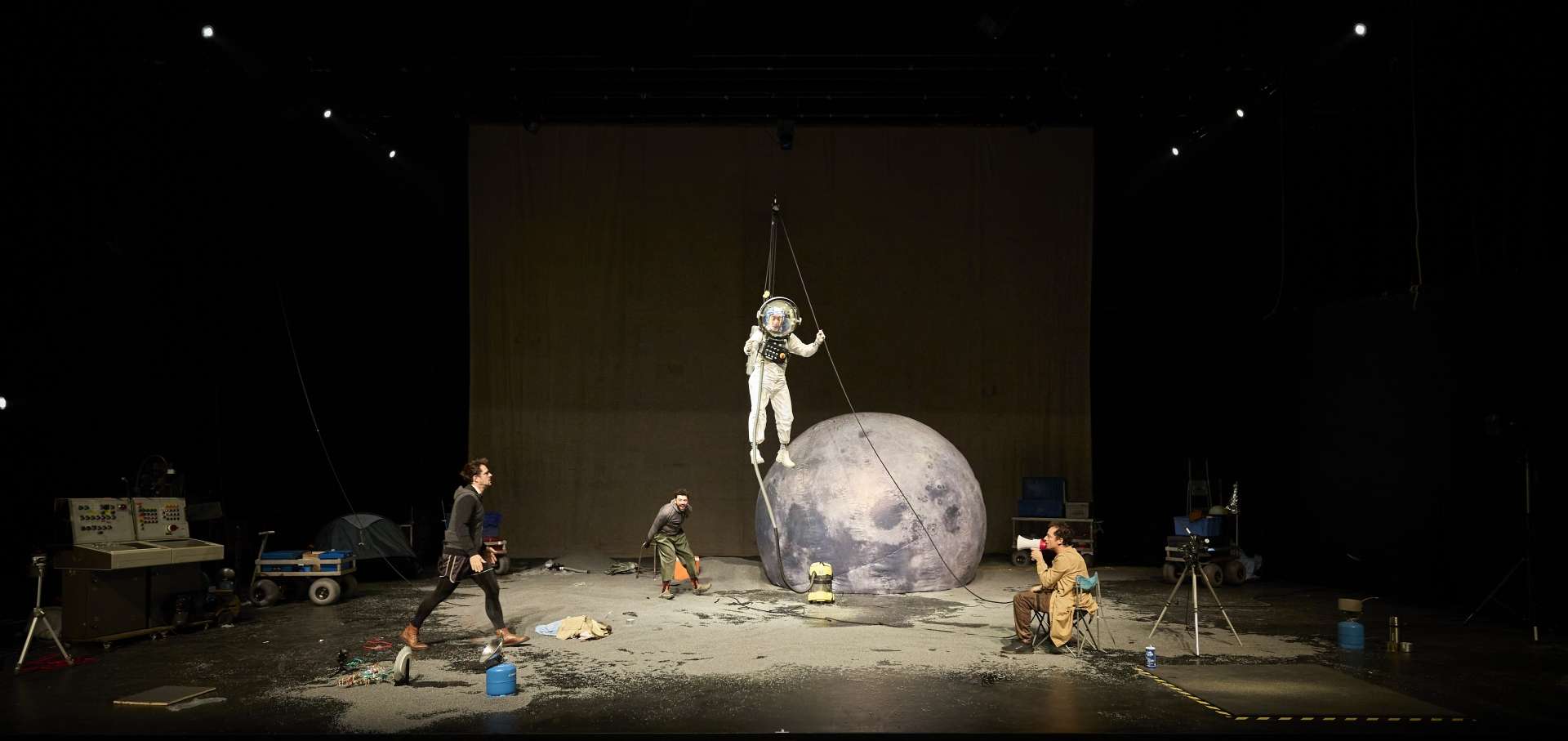 L'image montre une scène de (faux) premiers pas sur la Lune. On voit au centre, un astronaute suspendu en hauteur par des fils, à côté de lui, une grande sphère gonflable ressemblant à la Lune. Plusieurs personnes en costume, travaillant autour de l'astronaute. L'arrière-plan est composé d'un grand rideau.