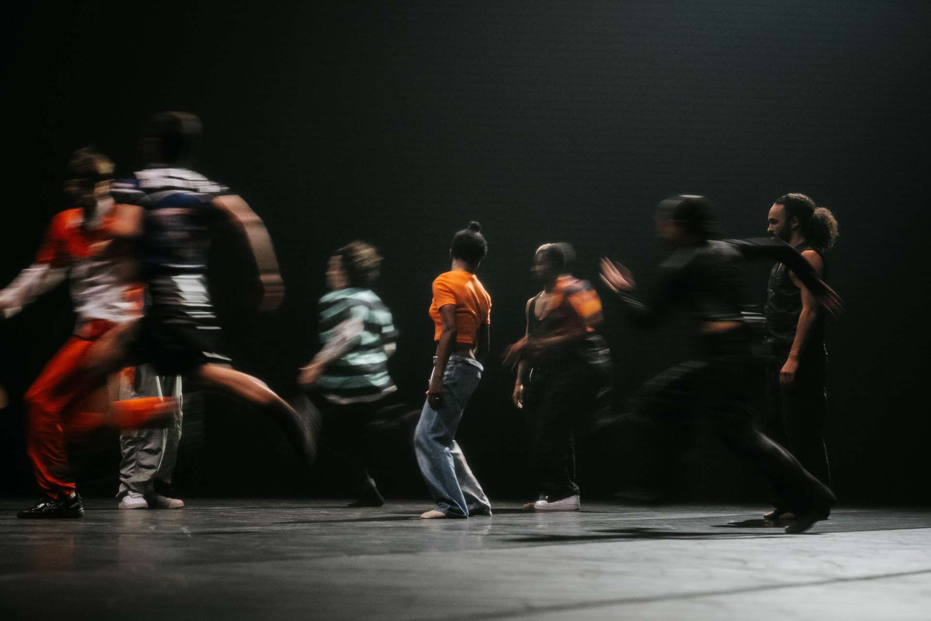 Photo d'un groupe de danseurs, danseuses, acrobates en pleine course sur la scène. Les mouvements sont flous. Seule une jeune femme, habillée d'un t-shirt orange fluo, se tenant debout au centre de l'image est nette et immobile.