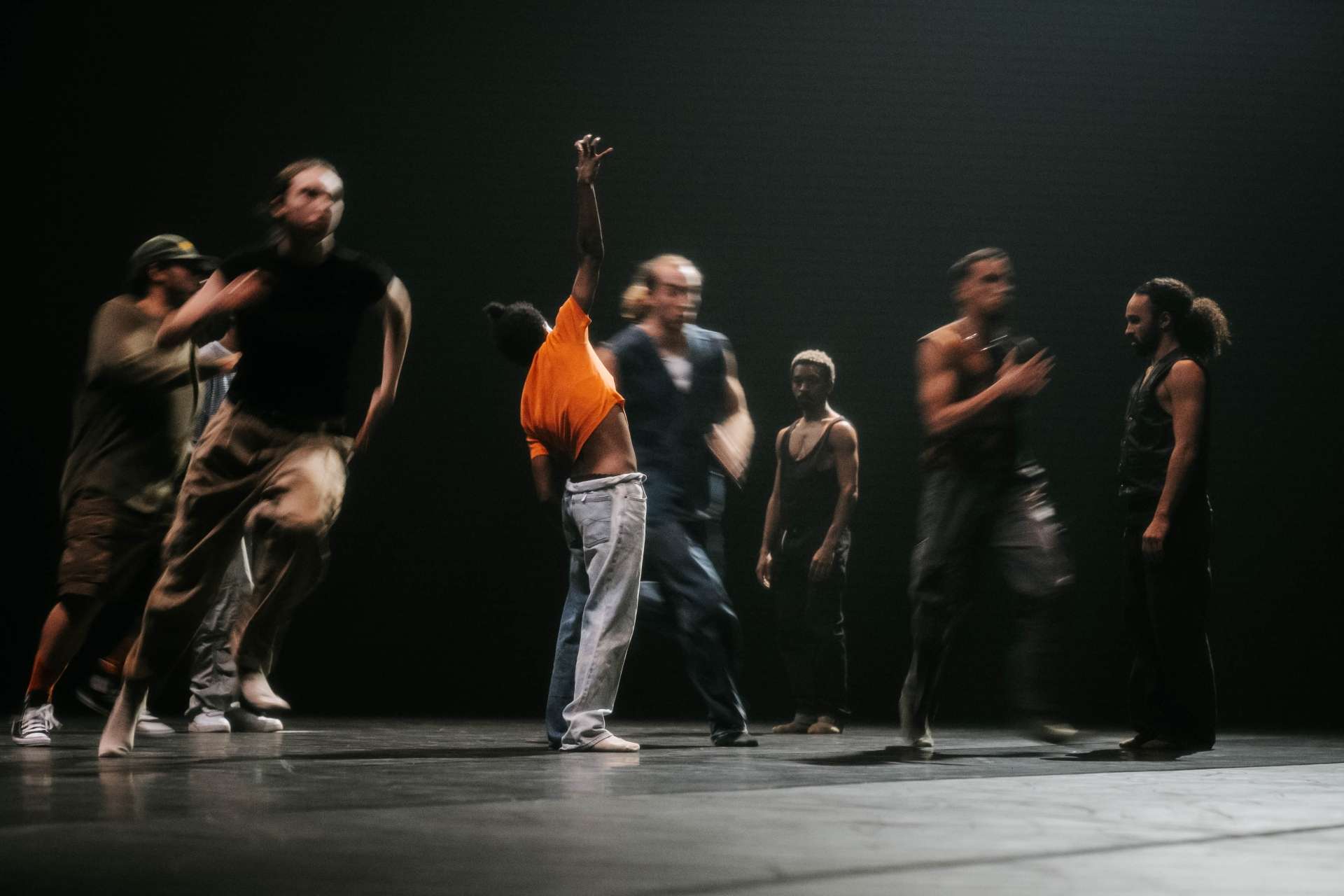 Photo d'un groupe de danseurs, danseuses, acrobates en pleine course sur la scène. Les mouvements sont flous. Seule une jeune femme, au centre de l'image, habillée d'un t-shirt orange fluo, se tenant debout avec un bras levé est nette.