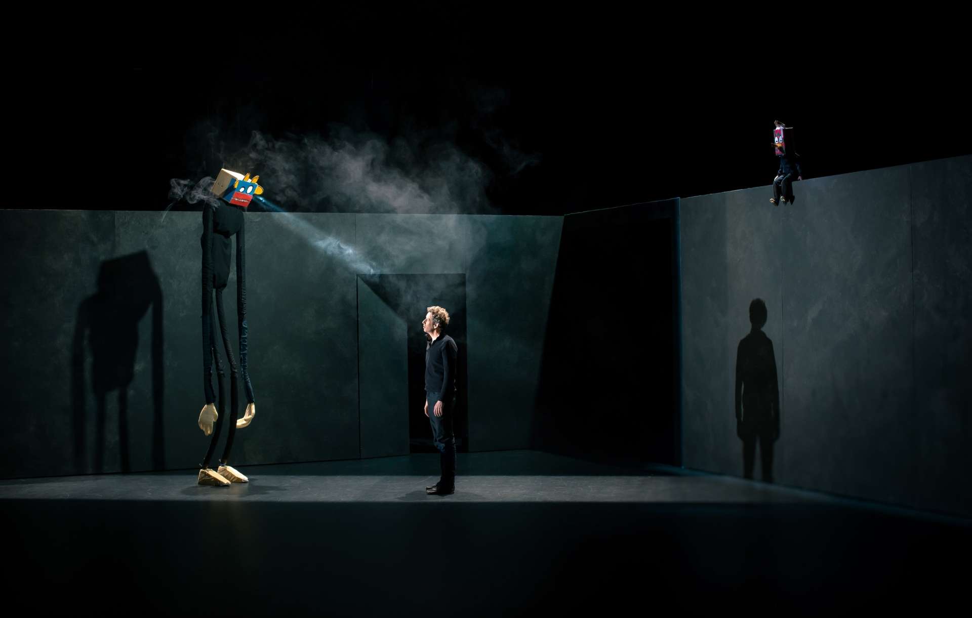 3 personnages sont présents sur cette image : un homme, de dos, en position debout, habillé d'un vêtement noir, regardant vers une source de lumière légèrement au-dessus de lui. Une géante marionnette, à gauche, a un corps et des membres longs et fins, avec une boîte colorée sur sa tête, ainsi que de longues ombres. Une figure avec un masque est assise sur le mur de droite.