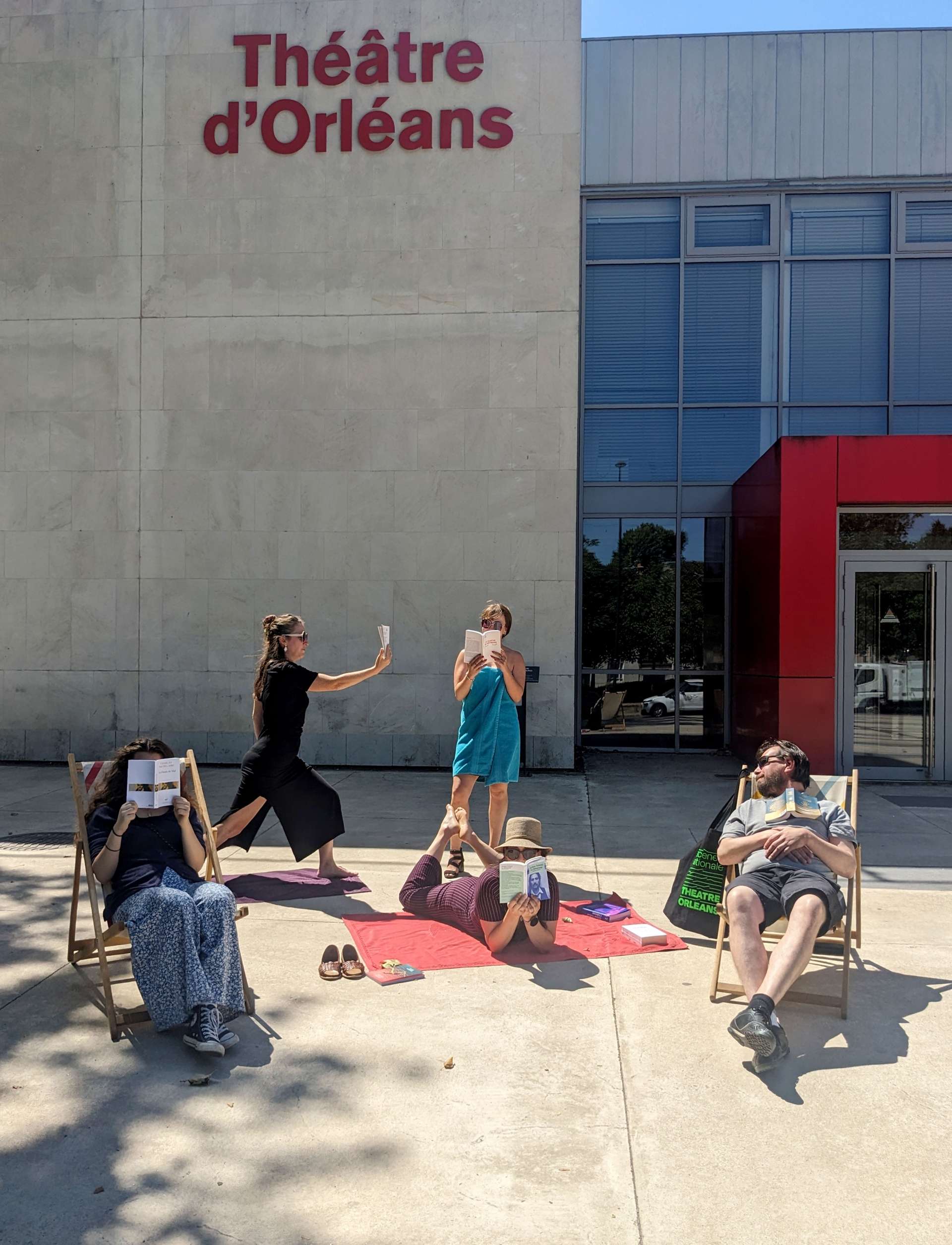 Devant la façade du Théâtre, sur des transats ou même en serviette sur le sol, cinq personnes de l'équipe de la Scène nationale sont en train de bouquiner. 