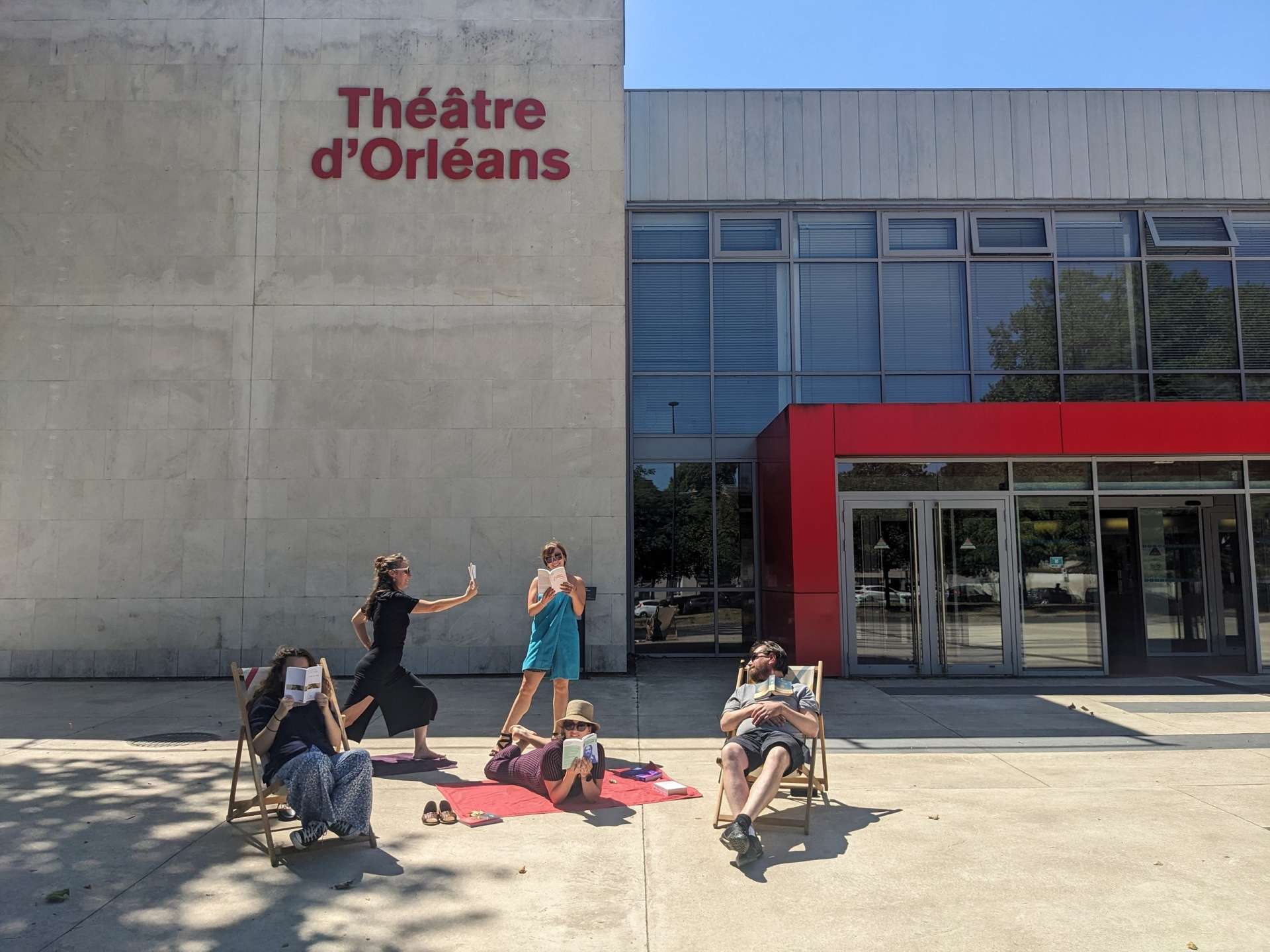 Devant la façade du Théâtre, sur des transats ou même en serviette sur le sol, cinq personnes de l'équipe de la Scène nationale sont en train de bouquiner. 