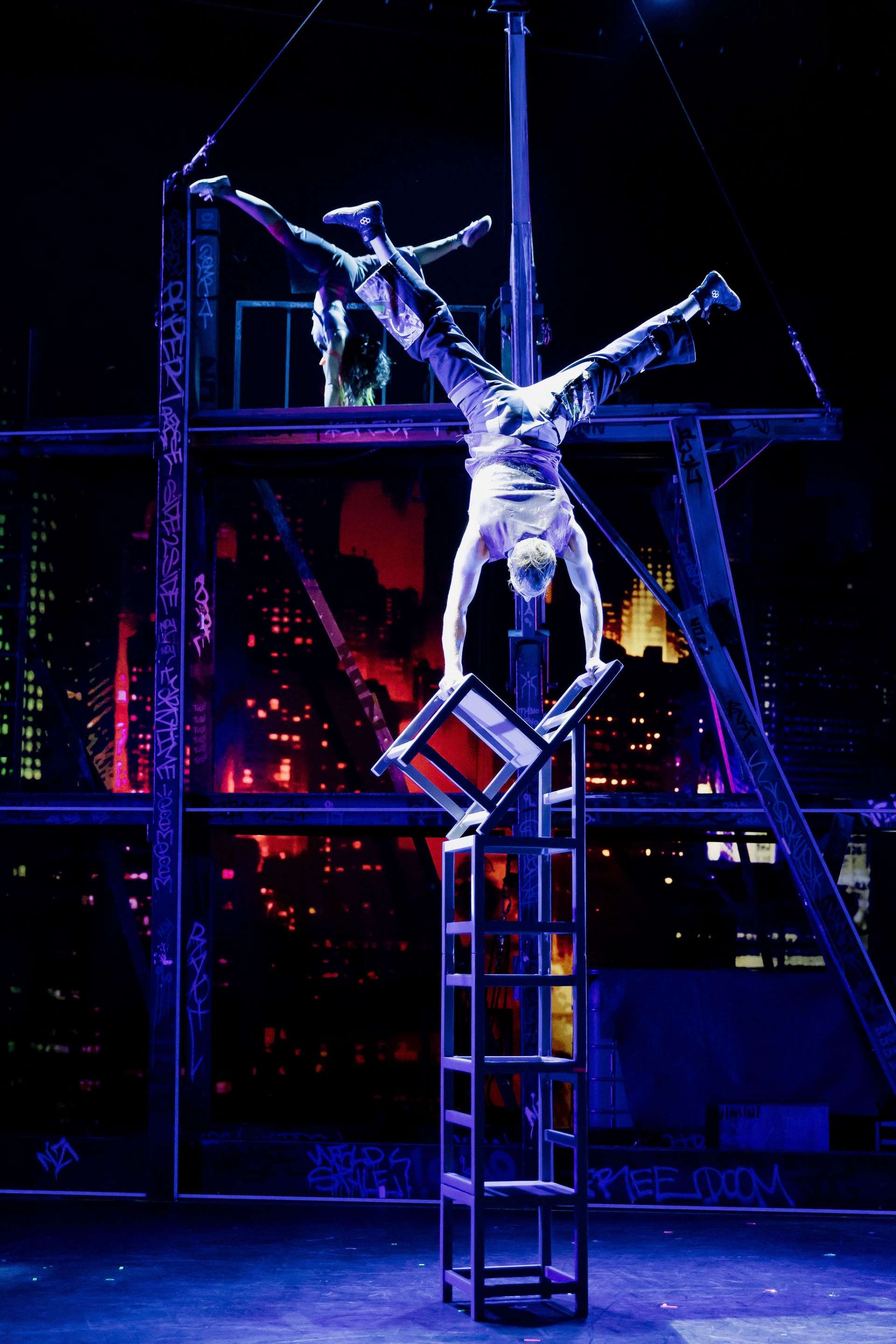 Un homme torse nu est retourné, tête en bas, sur un support en bois. Le décor au fond ressemble à une projection vidéo. 