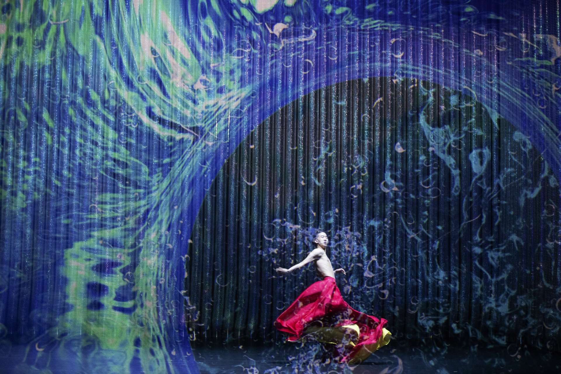 Au premier plan, un danseur danse torse nu et en robe rouge. Il est enveloppé d'une projection vidéo grandiose avec des teintes bleues. Cela ressemble à une vague. 
