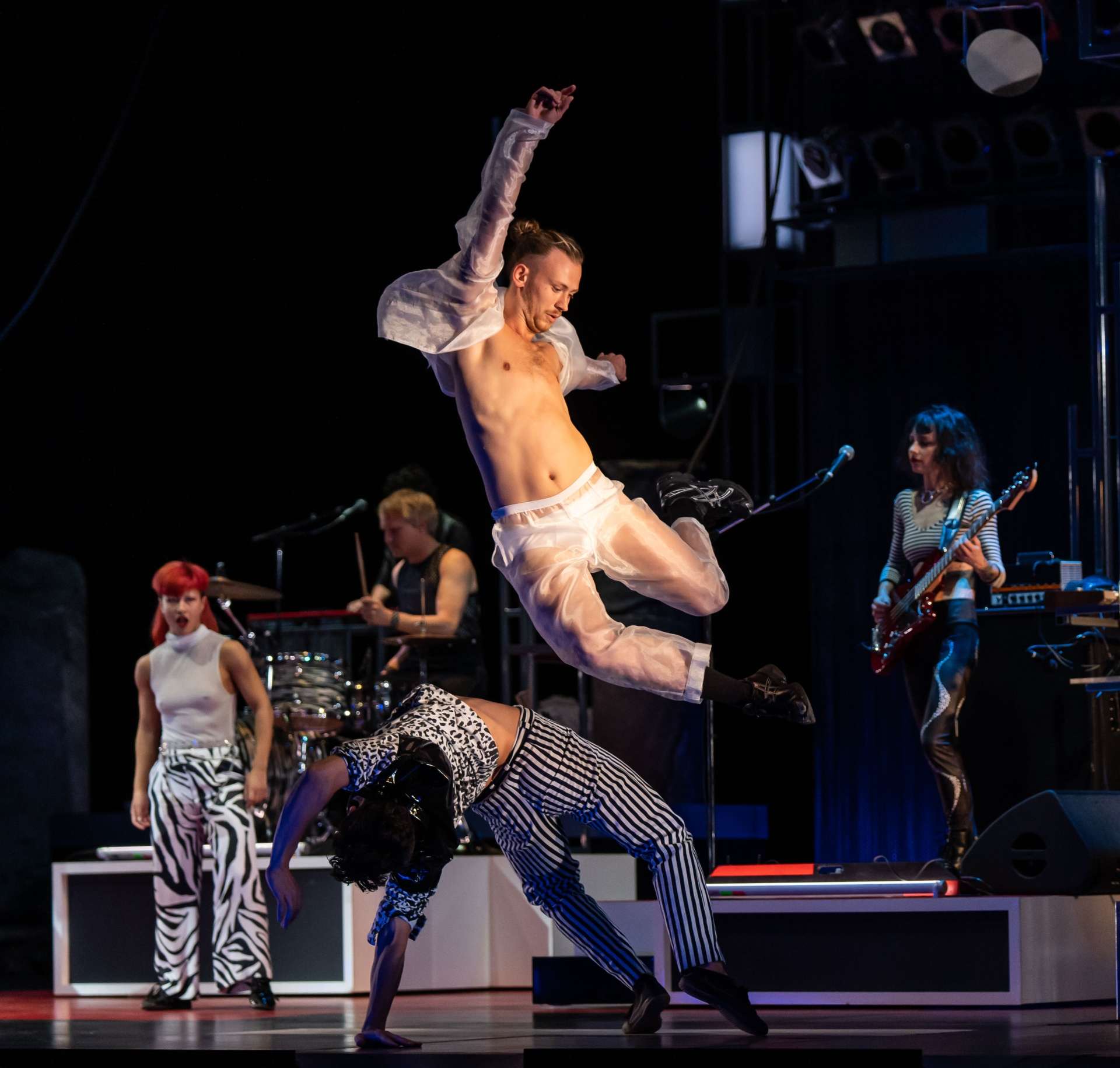 Un danseur vêtu d'un costume transparent blanc est en plein saut. En fond de scène un autre interprète effectue une figure au sol devant le trio rock qui accompagne la pièce. 