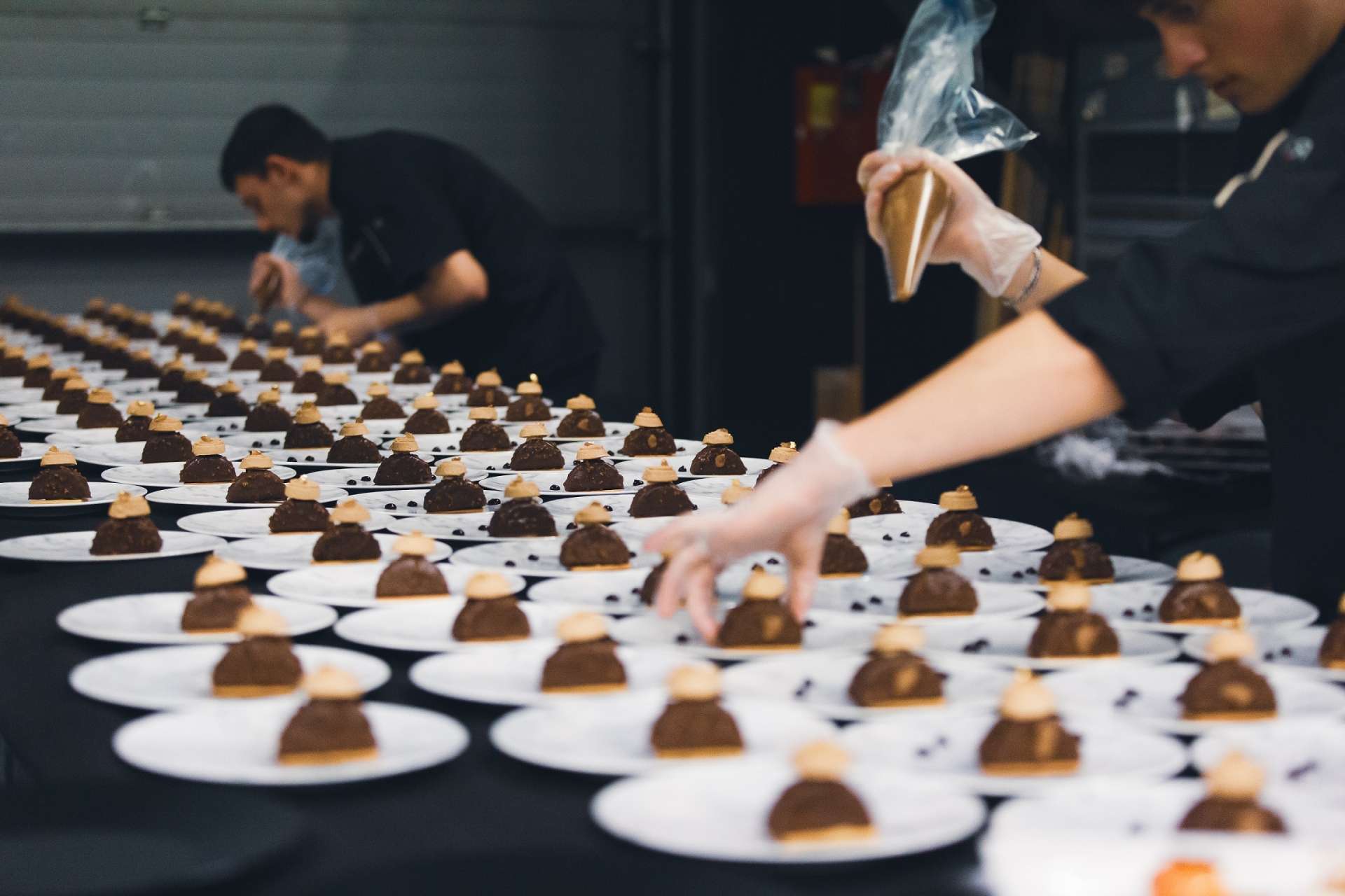 Une multitude d'assiettes sont alignées. Deux personnes s'affairent à mettre de la crème sur des desserts. 