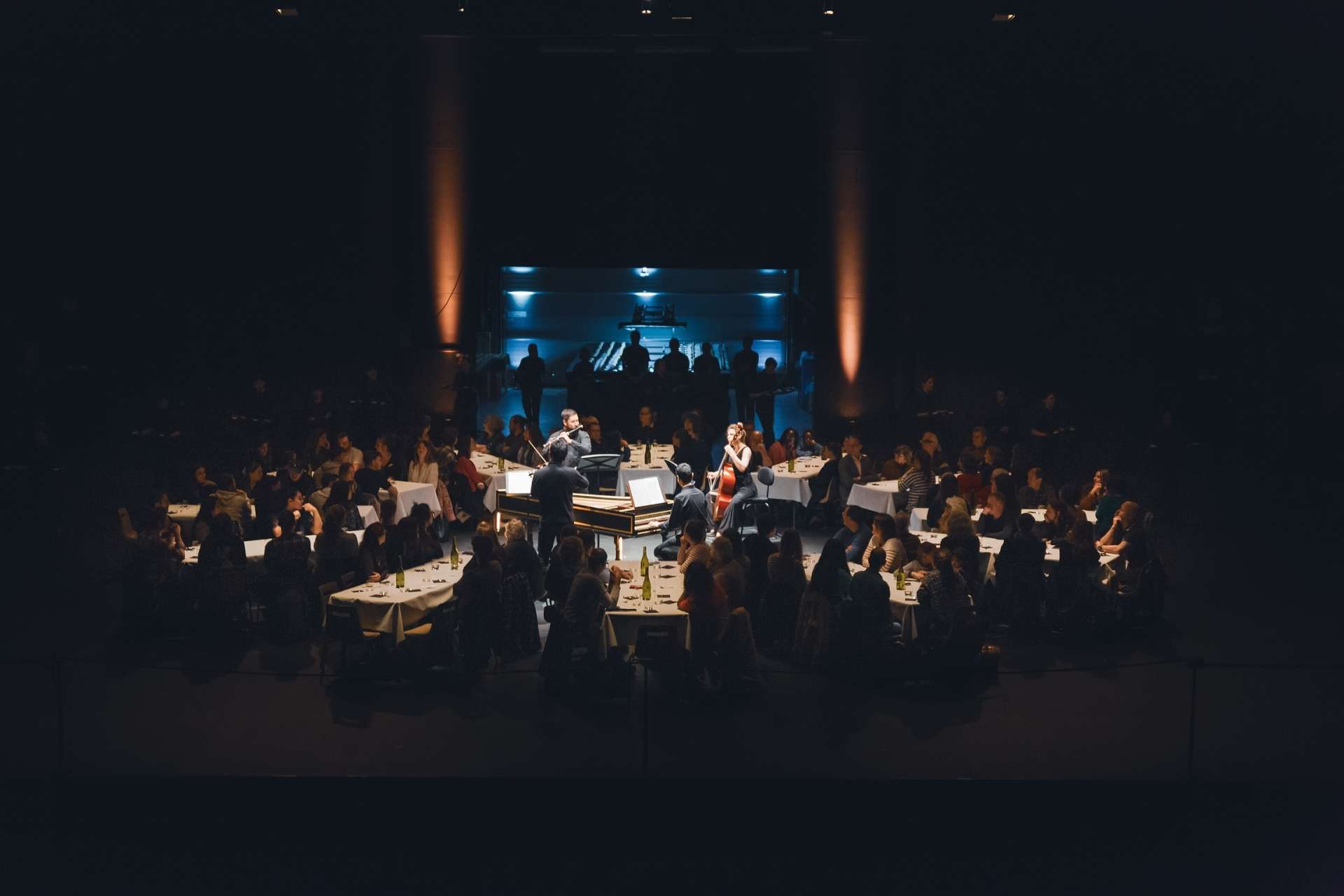 Plan large où l'on aperçoit les convives installés à leurs tables et regardant les musiciens au centre. Les pâtissiers sont visibles en fond de salle. Il y a une jolie ambiance lumineuse. 