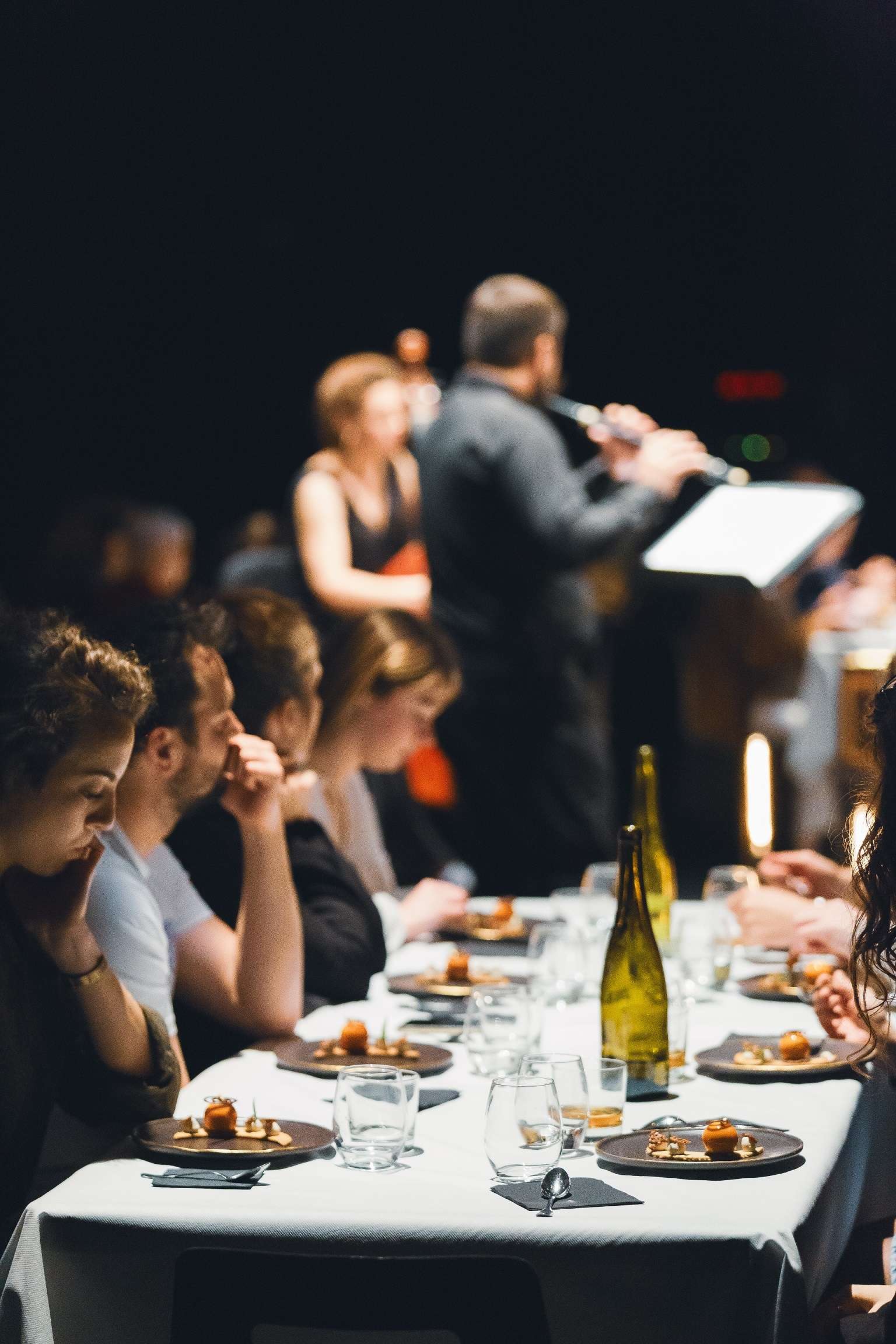 Une table avec des convives, des assiettes avec des desserts et un peu plus loin au centre, des musiciens. 