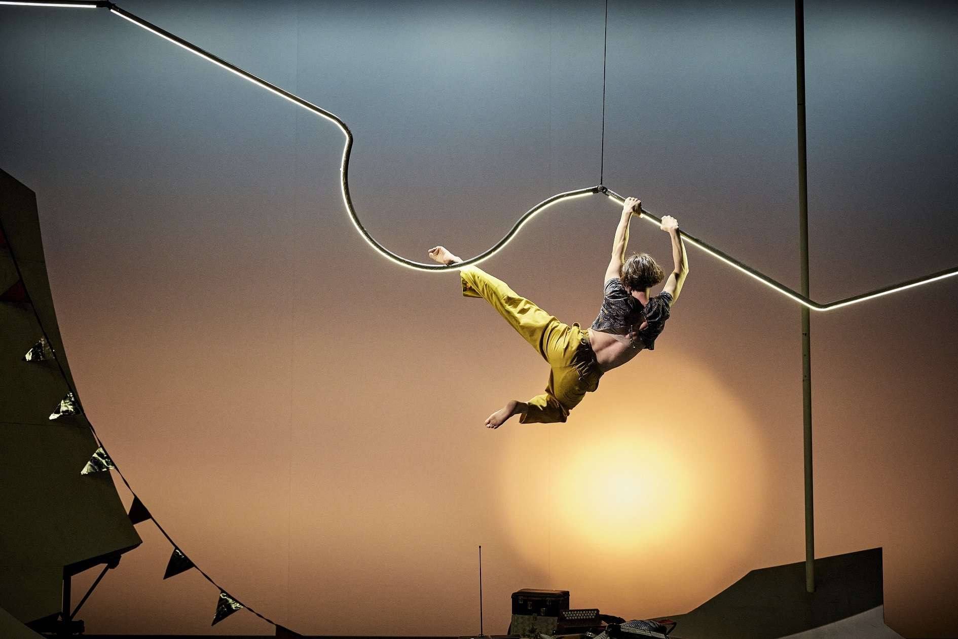 Dans une lumière digne d'un beau couché de soleil, une acrobate est suspendue dans les airs, accrochée à une barre métallique qui ondule. 