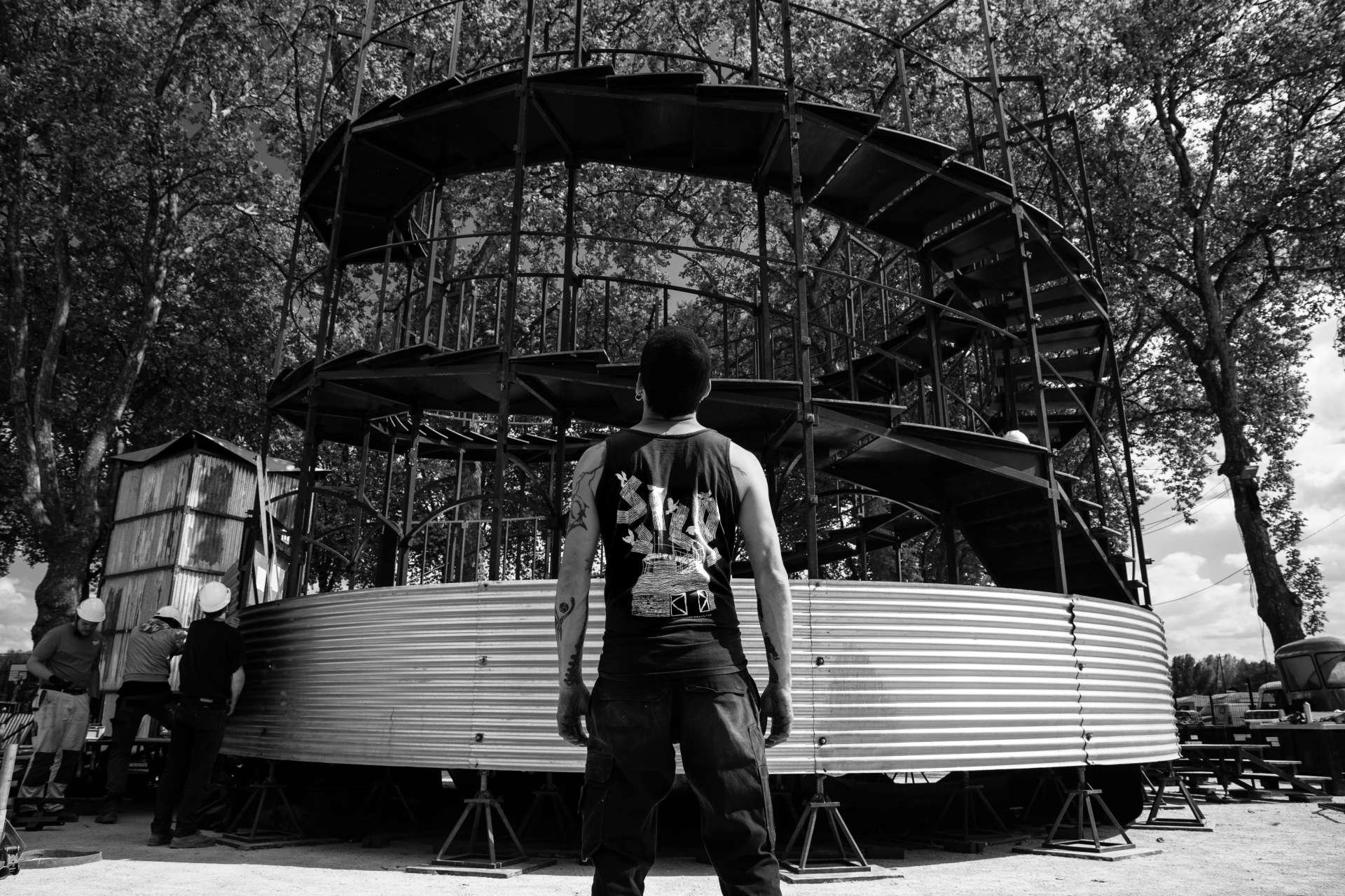 Photo monochrome du montage du silo pour L'Absolu, aperçu d'un technicien en premier plan avec un débardeur "Le silo" devant la structure. 