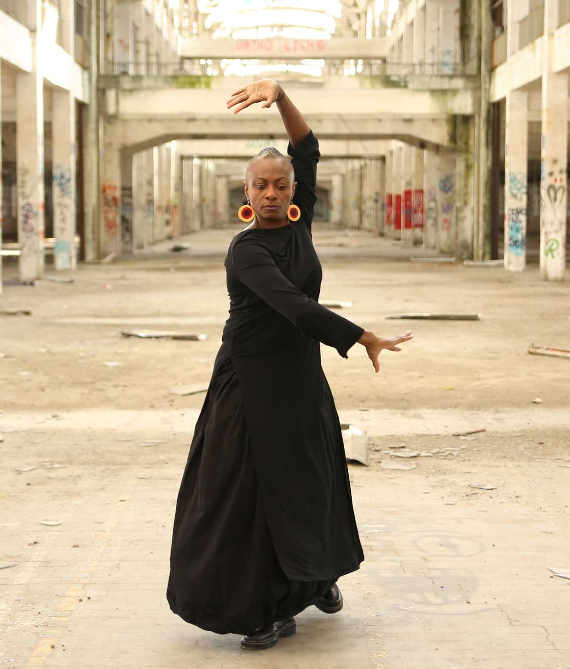 Sur fond de friche industrielle, la danseuse Sophiatou Kossoko pose au premier plan. Elle est habillée d'une grande robe noire qui contraste avec le fond l'ambiance beige grise de l'entrepôt.