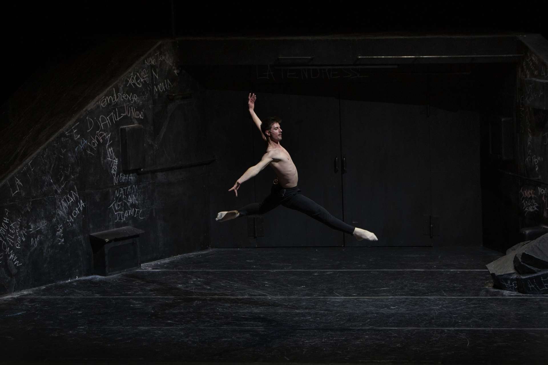 Sur scène, dans un décor noir, avec des murs au fond recouverts d’écritures à la craie blanche, un jeune danseur classique torse nu à la peau blanche, avec un leggin noir, fait un grand jeté.