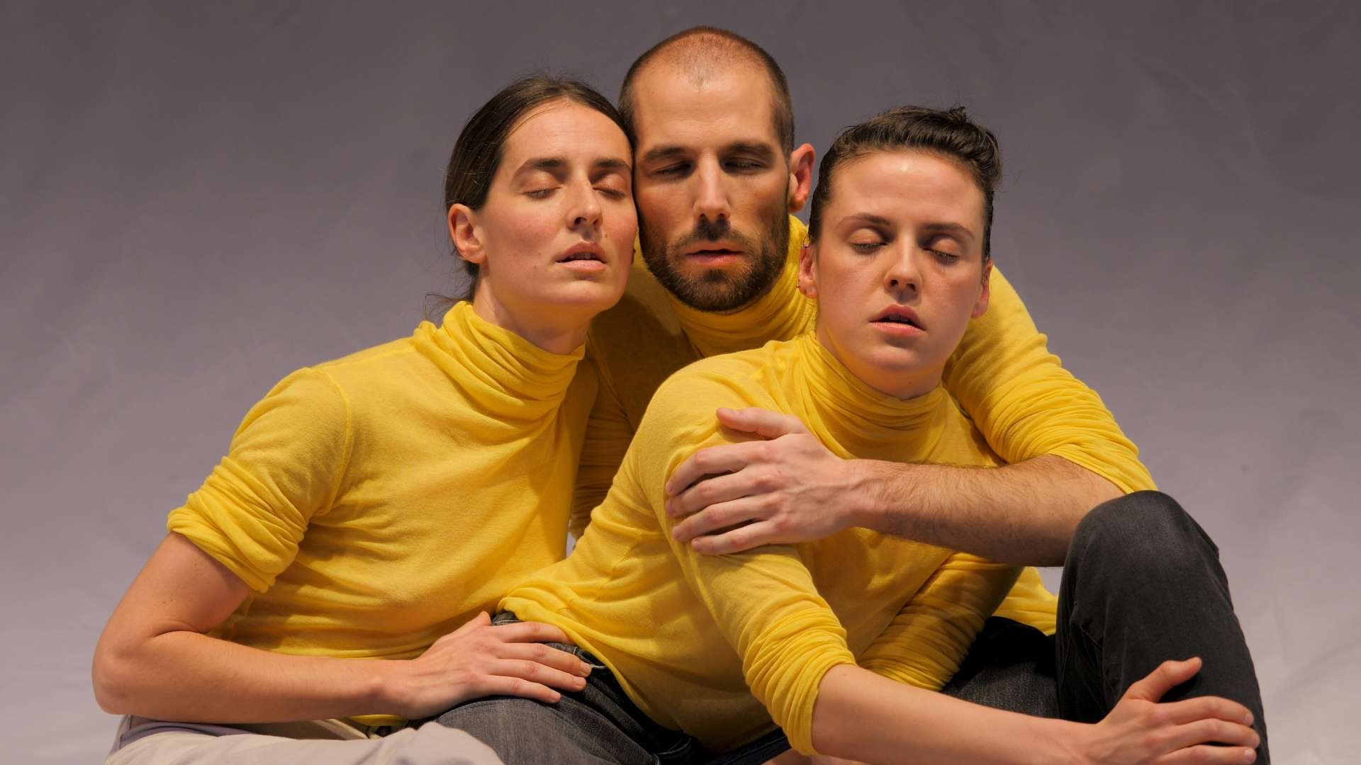 Deux danseuses et un danseur, vêtus à l’identique d’un pull-over à col roulé jaune, se tiennent enlacés, les yeux fermés.