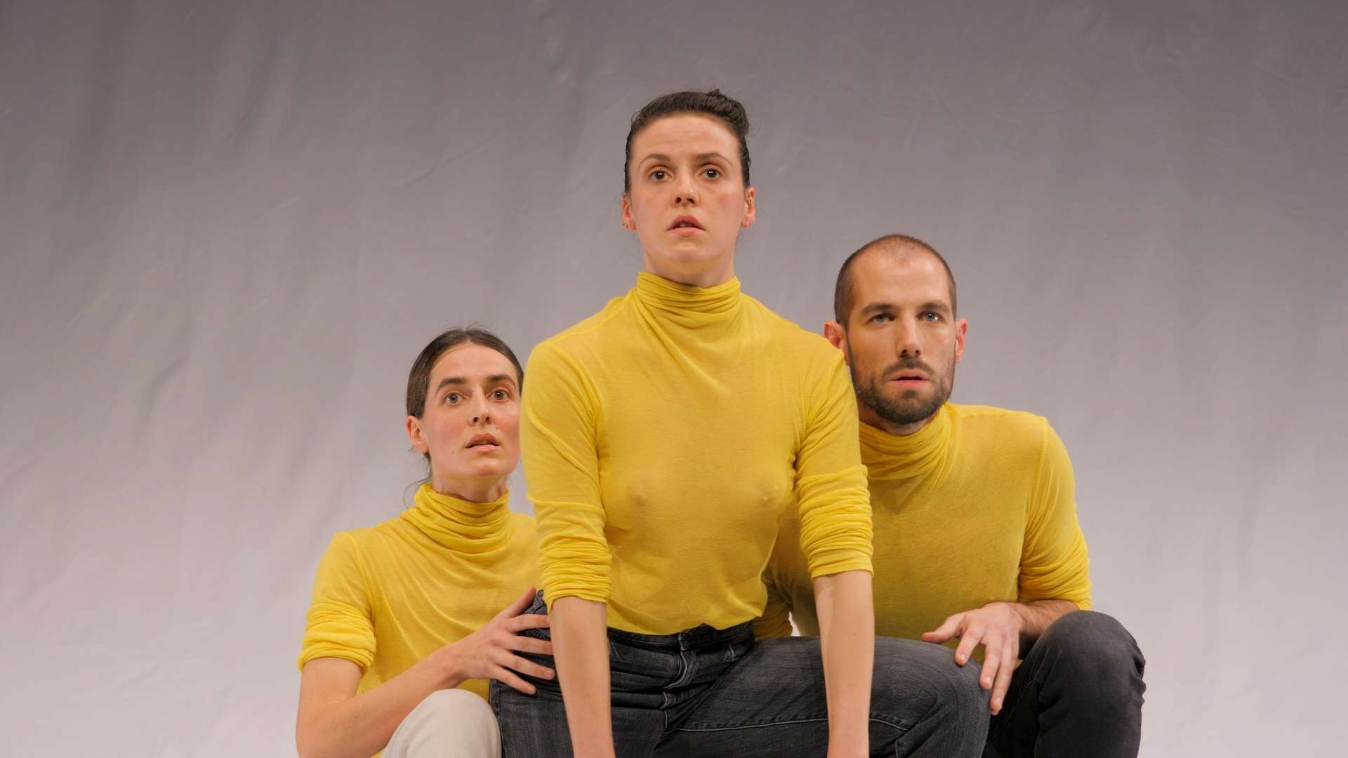 Deux danseuses et un danseur, vêtus à l’identique d’un pull-over à col roulé jaune, se tiennent debout dans une posture chorégraphique.