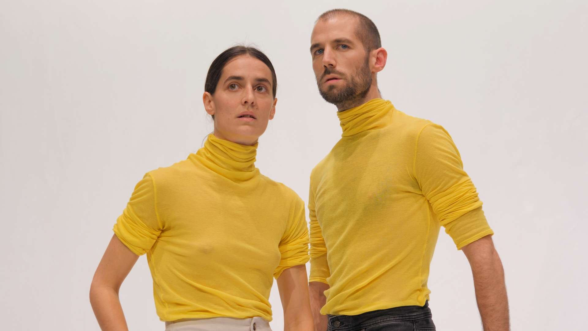 Un danseur et une danseuse, vêtus de pulls à col roulé jaunes assortis, se tiennent côte à côte, le regard tourné dans la même direction